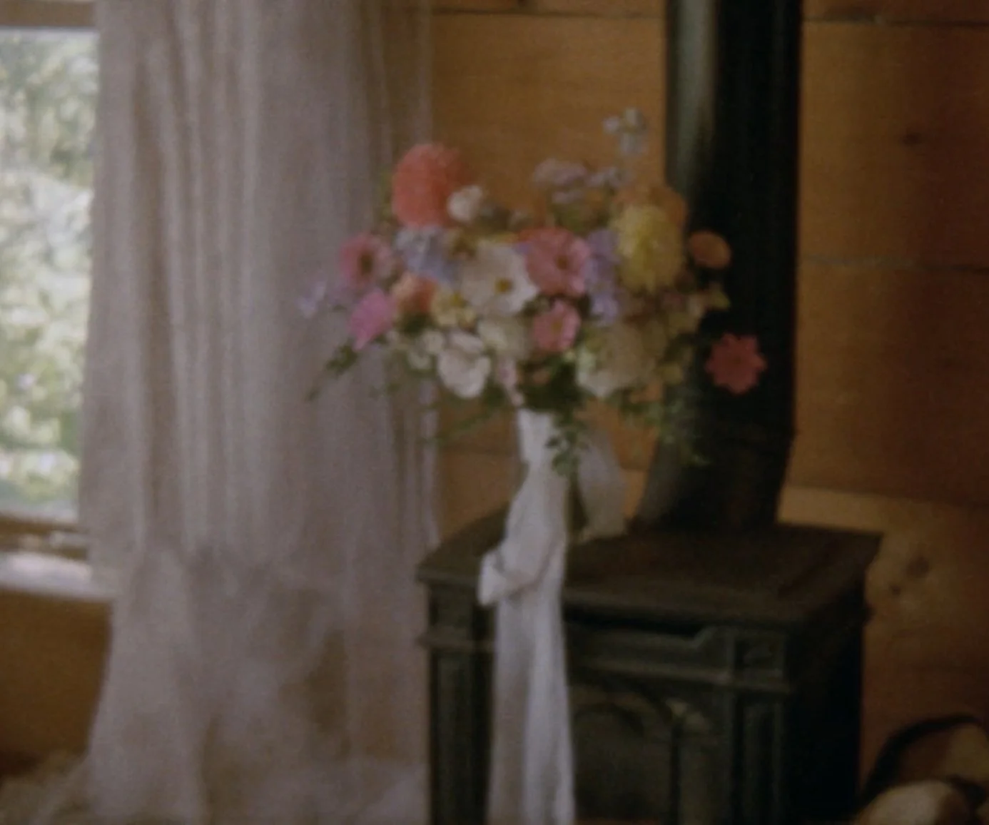 A floral wedding bouquet on a table in a cozy wooden room with a window and curtains.