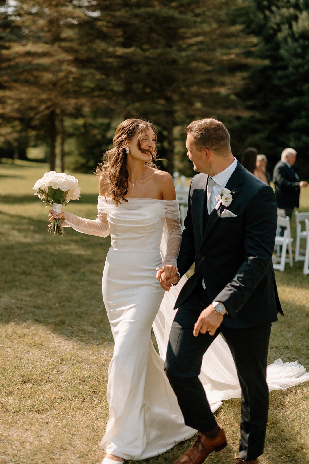 Bride and groom holding hands, smiling, walking outdoors during wedding ceremony at Cox Creek Cellars, bride in white dress, groom in black suit, natural background.