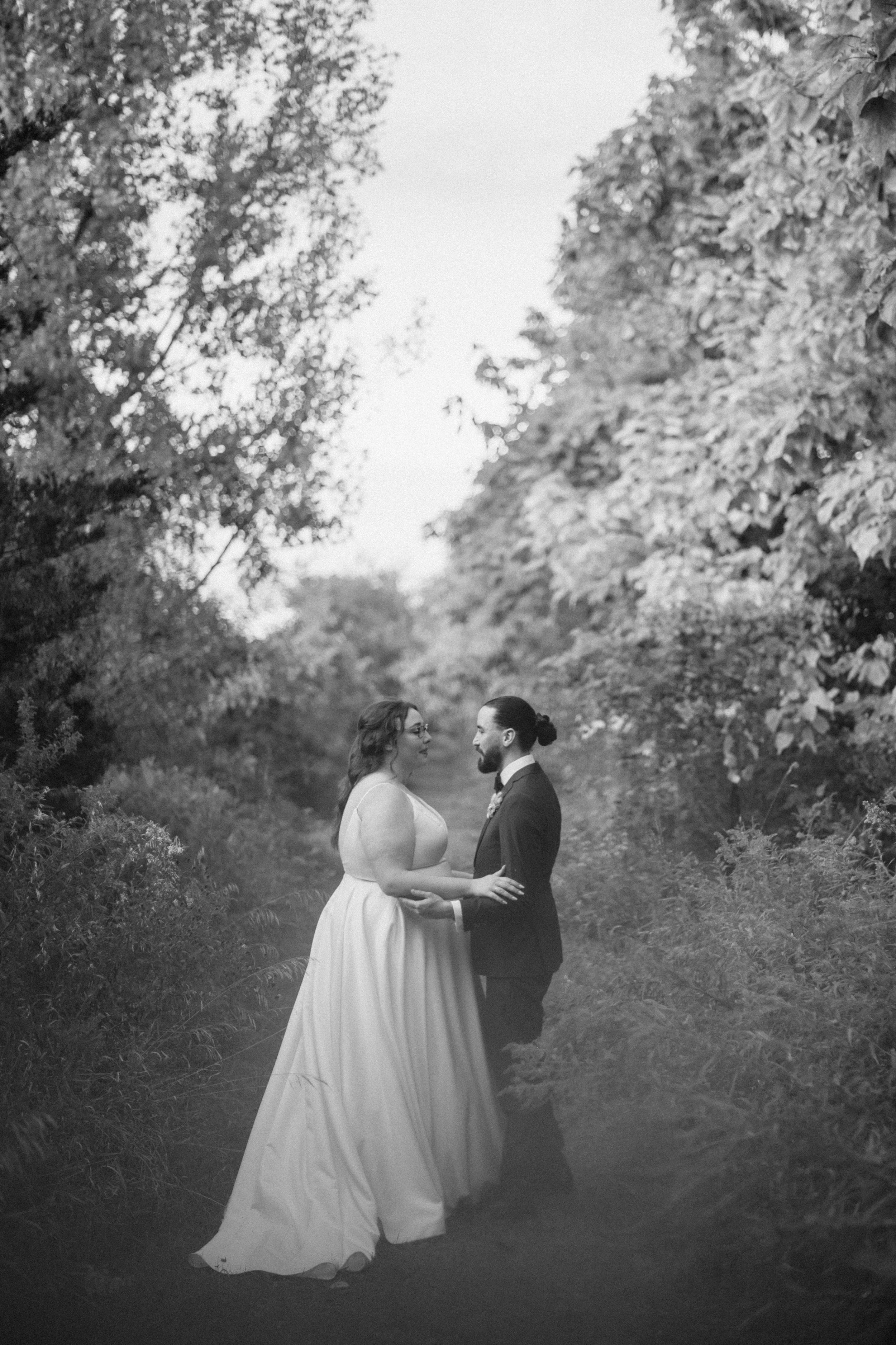 A black and white photo of a couple in wedding attire holding hands and looking at each other in a wooded outdoor setting, 100 Acre Wood.
