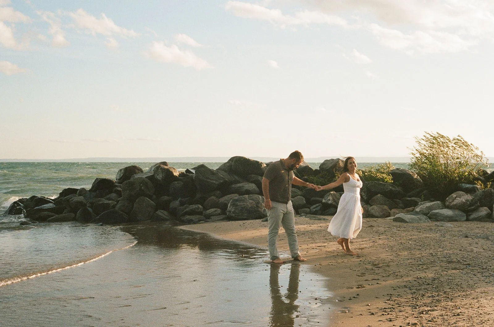 A couple holding hands and walking barefoot along the shoreline of Georgian Bay during an engagement session at sunset, with rocks and a bush in the background.