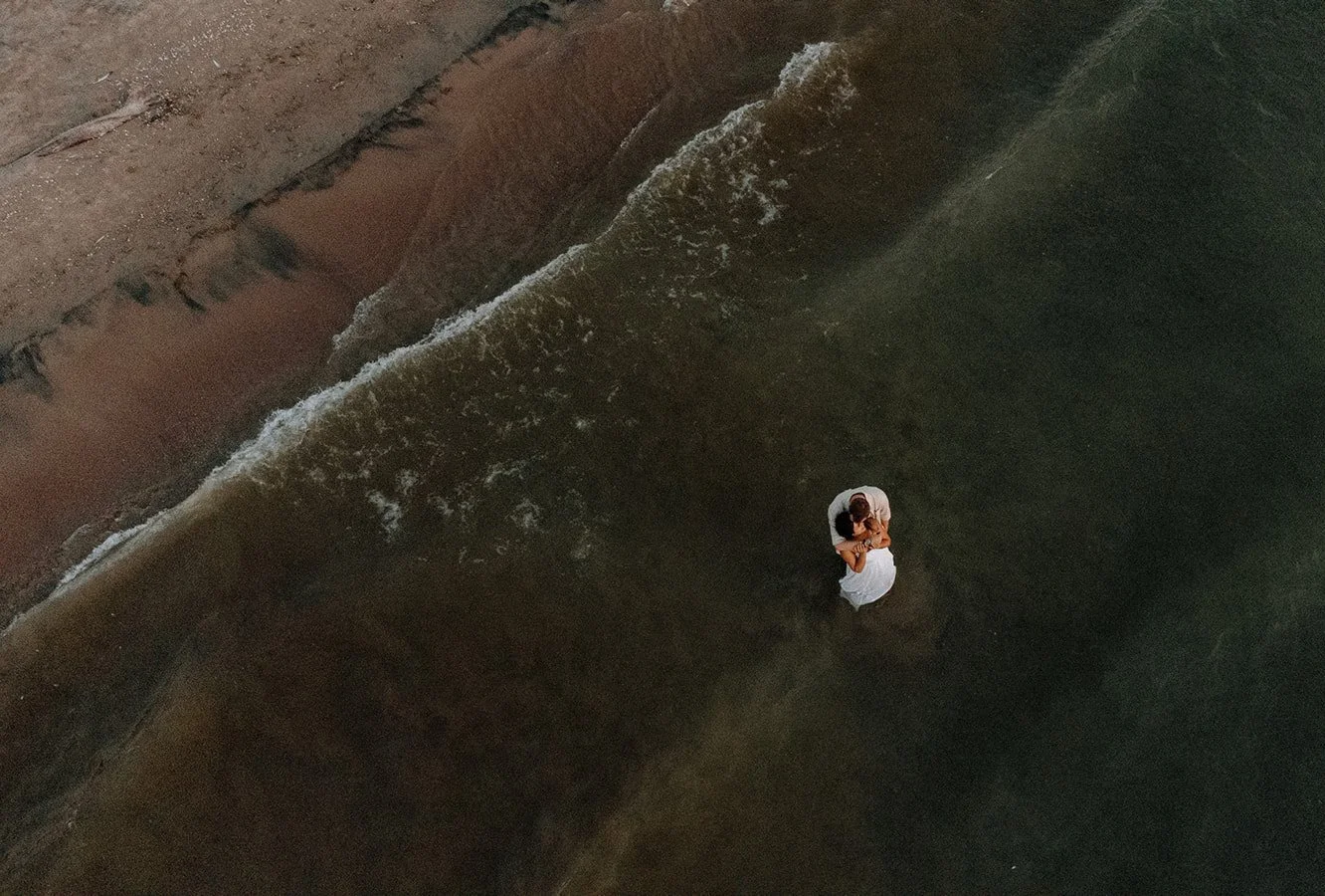 A couple standing close together in shallow water at the beach, near a sand barrier or wall.