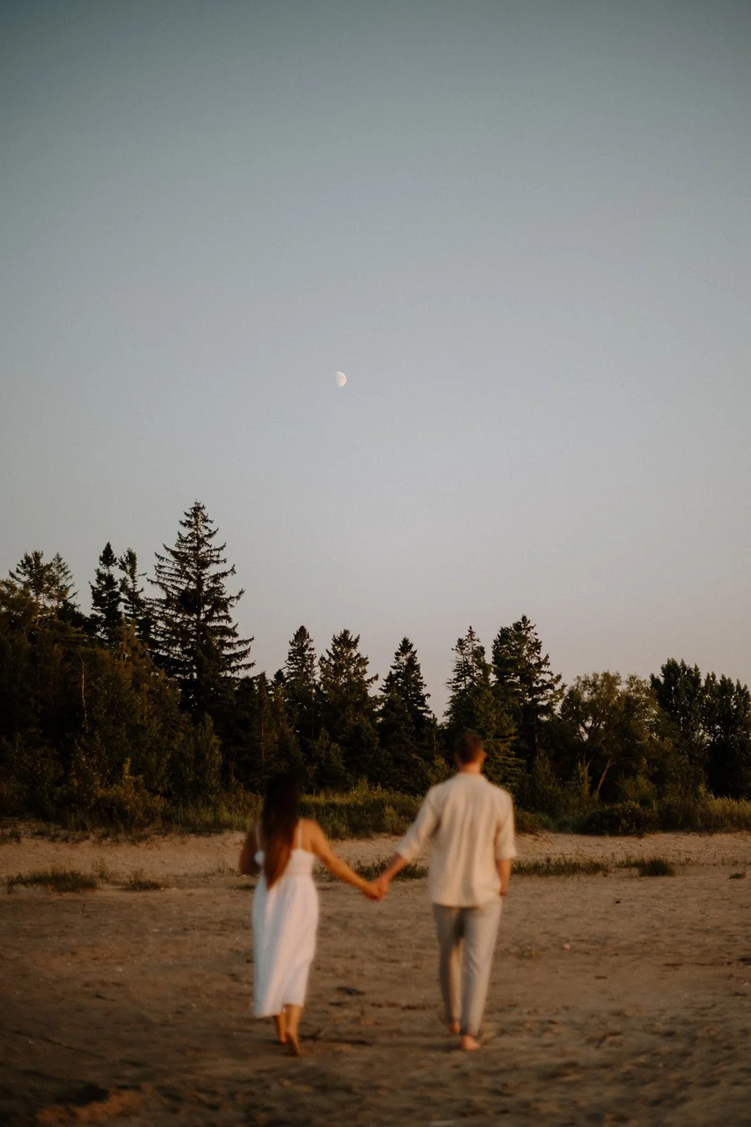 A couple holding hands walking on a sandy beach during sunset, with pine trees in the background and a visible moon in the sky.