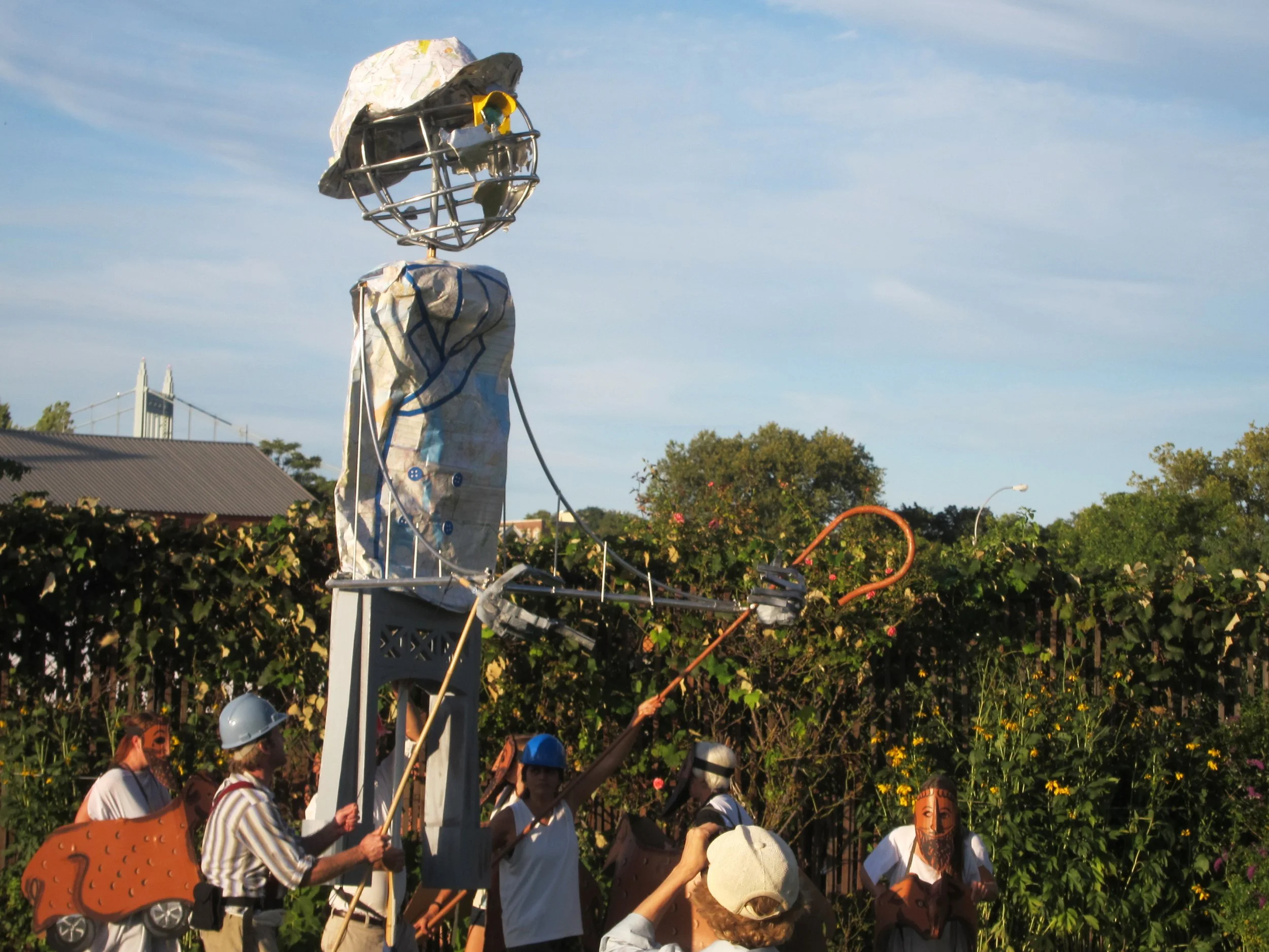A street art sculpture of a human figure with a globe as the head, made from various materials including wire, metal, and paper, is being installed or displayed outdoors among people wearing costumes, some with helmets, in a garden or park with trees and bushes in the background.
