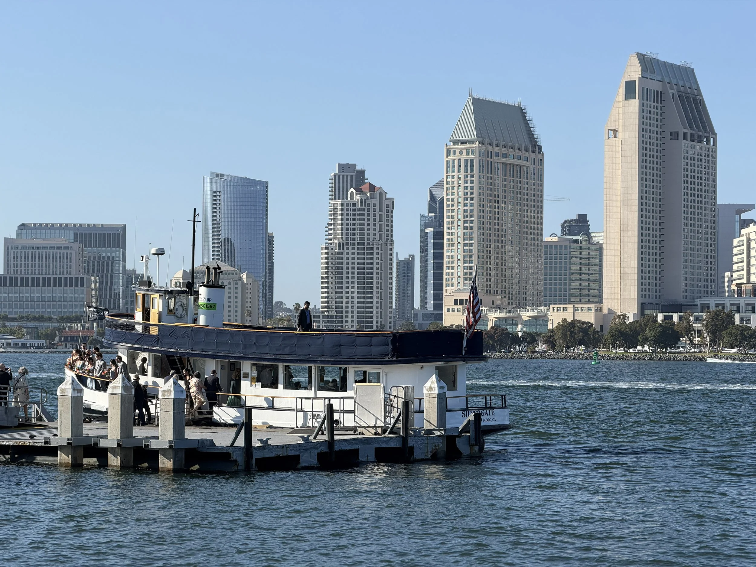 Photograph showing a passenger ferry docked at a pier with a city skyline in the background. The scene features calm water, clear blue sky, and several tall buildings, highlighting urban waterfront activity.