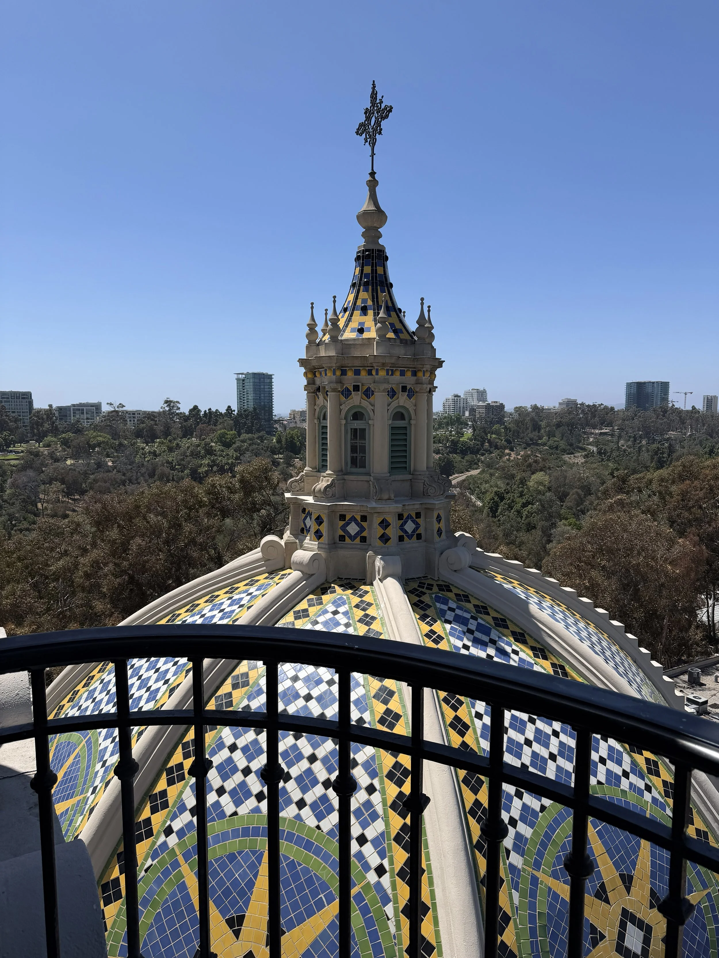 Photograph of a colorful tiled dome with intricate patterns in blue, yellow, white, and green, topped with a decorative finial and cross. The dome is viewed from behind a black metal railing, with a cityscape and clear blue sky in the background.