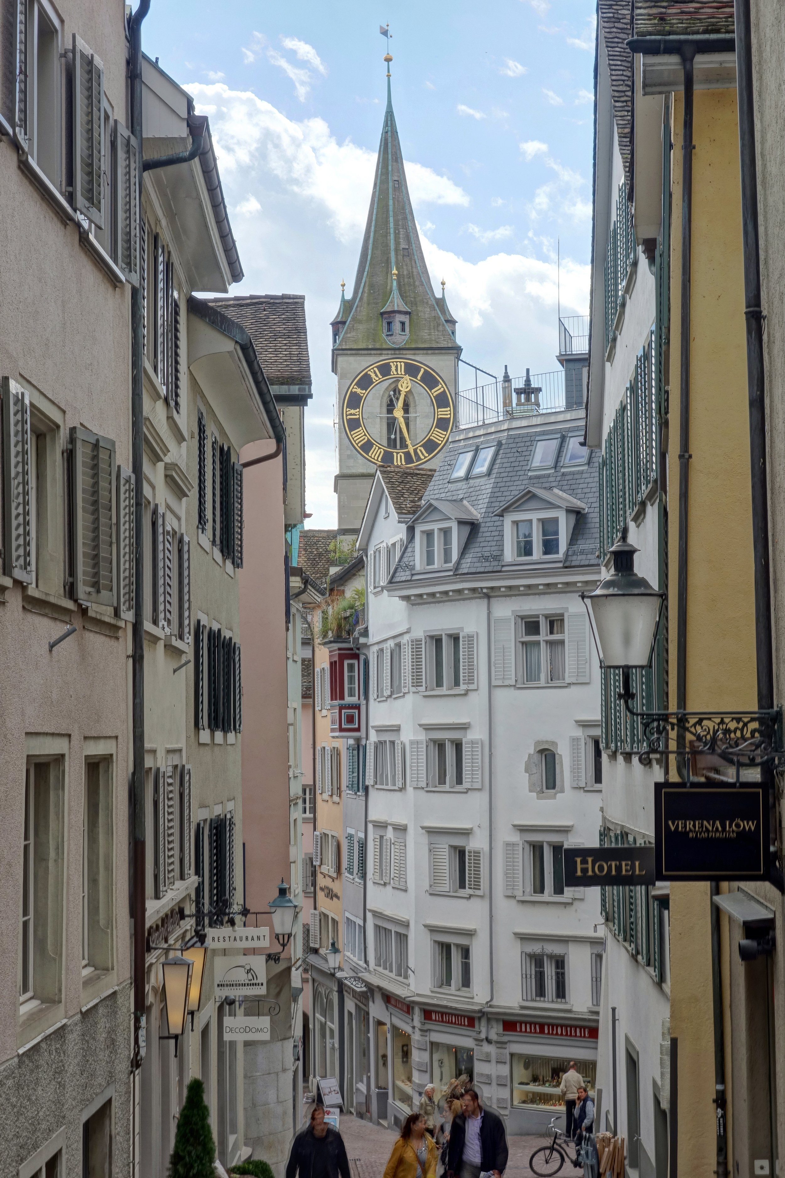 A narrow European street with multi-story buildings, some with open shutters, leading toward a clock tower with a tall spire in the background. Pedestrians walk along the cobblestone street, which features signs for hotels and shops.