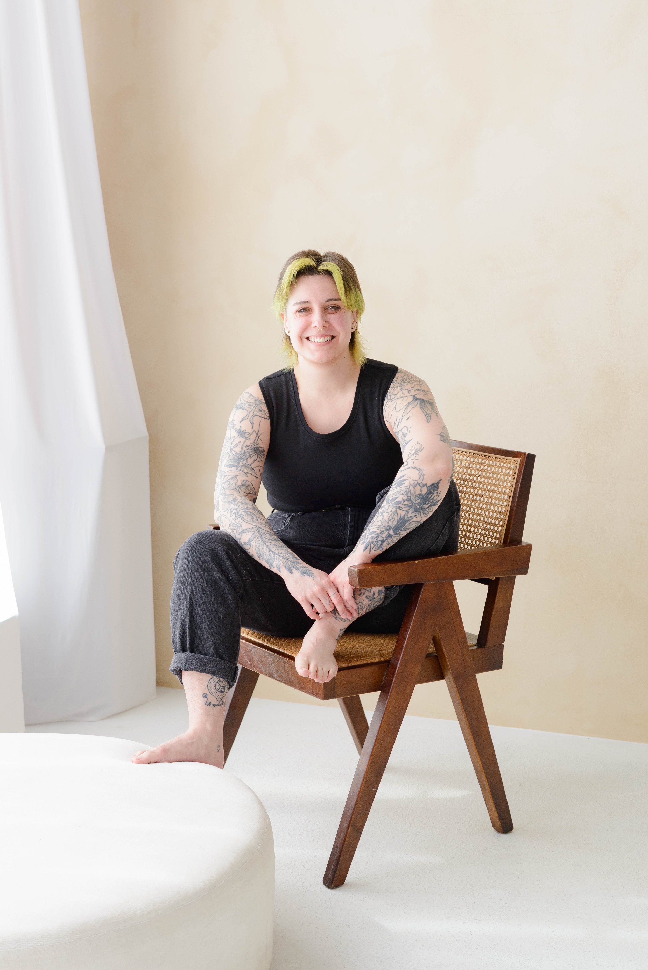 A smiling nonbinary person with short green hair and tattoos on their arms, wearing a black chest binder, sitting on a wooden chair with one leg crossed over the other, in a minimalistic room with beige walls and white curtain to the side.