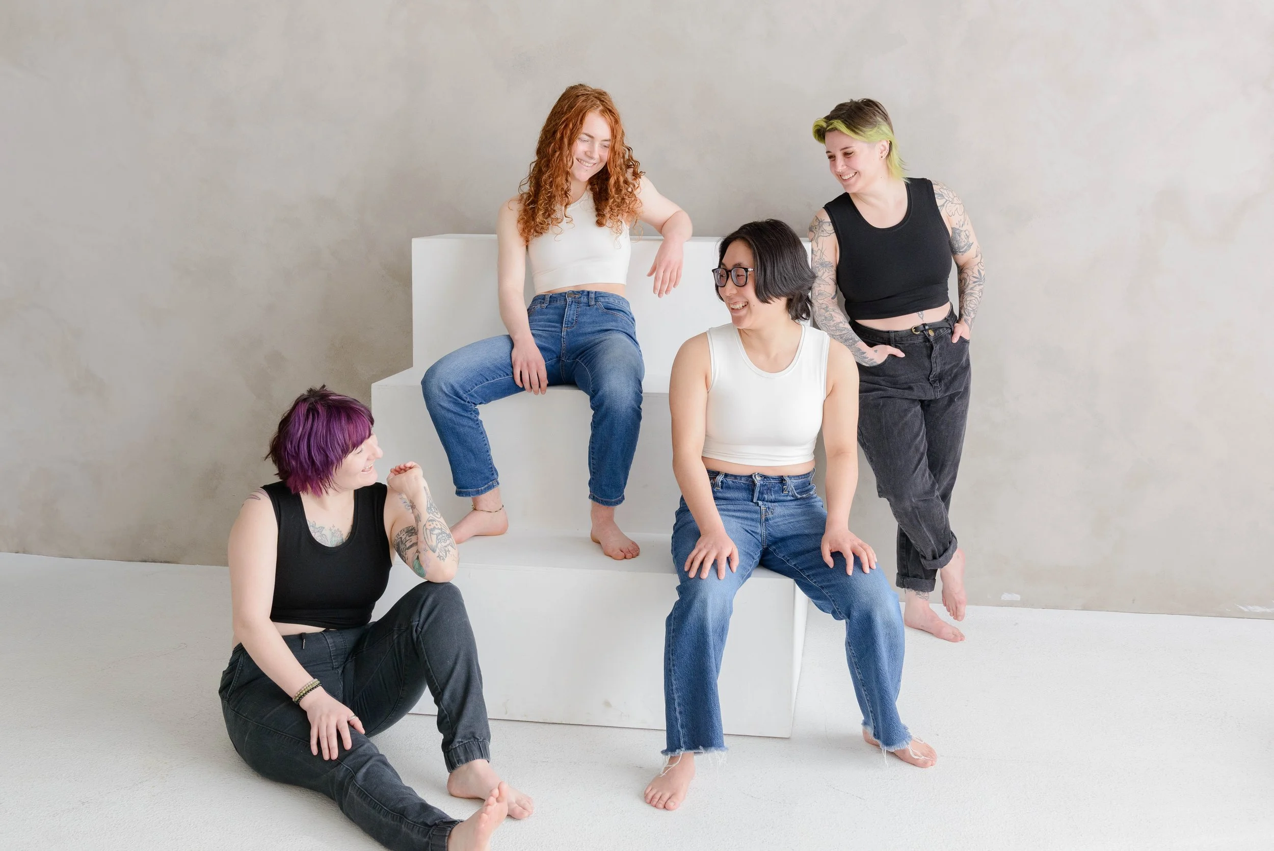 Four nonbinary people with tattoos and various hair colors, dressed in chest binders and jeans, are posing together in a studio with a plain background. They are sitting and standing on and around a white block, interacting and smiling at each other.