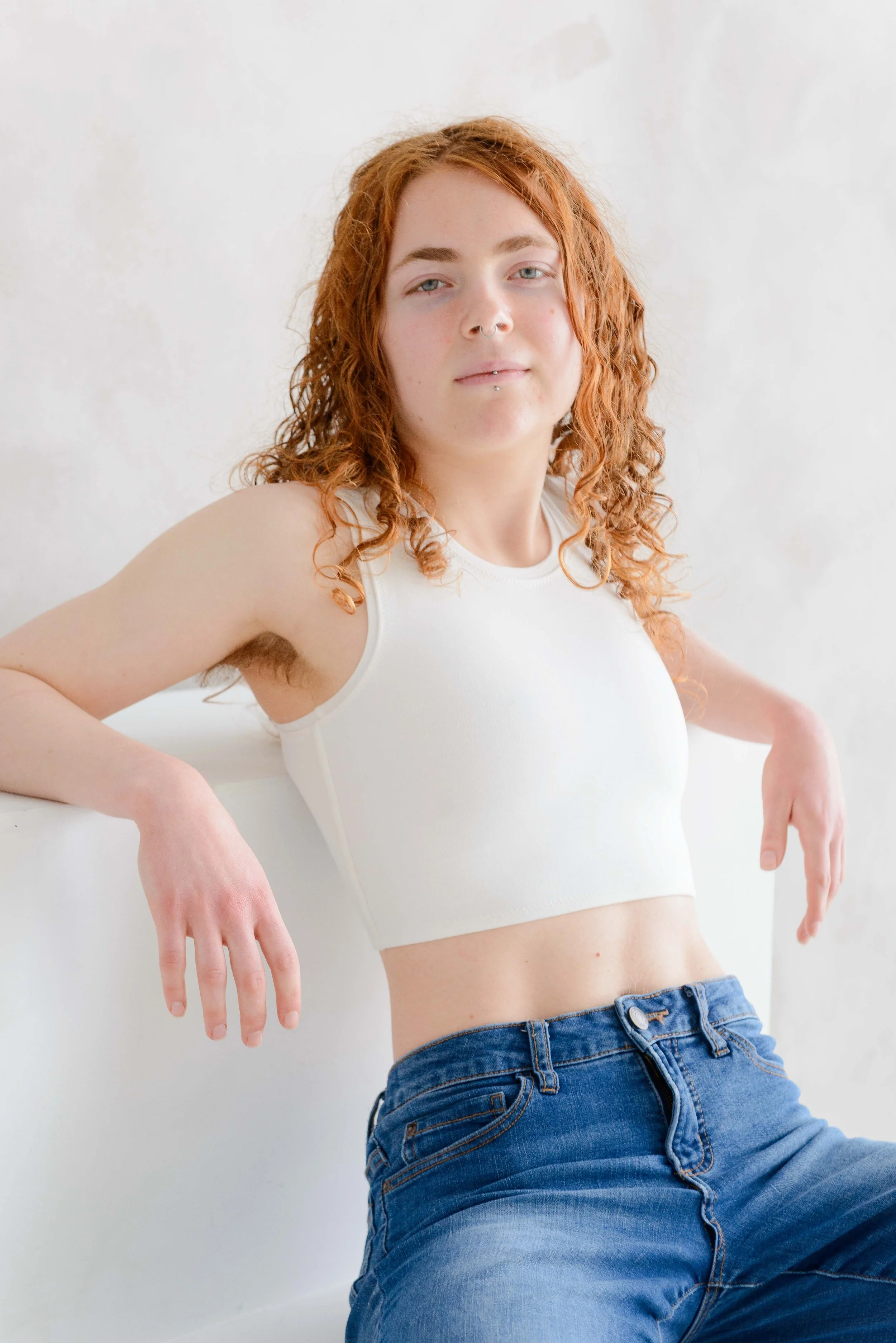 A person with curly red hair and light skin wearing a white chest binder and blue jeans, posing against a white wall with their hands and arms extended outward.