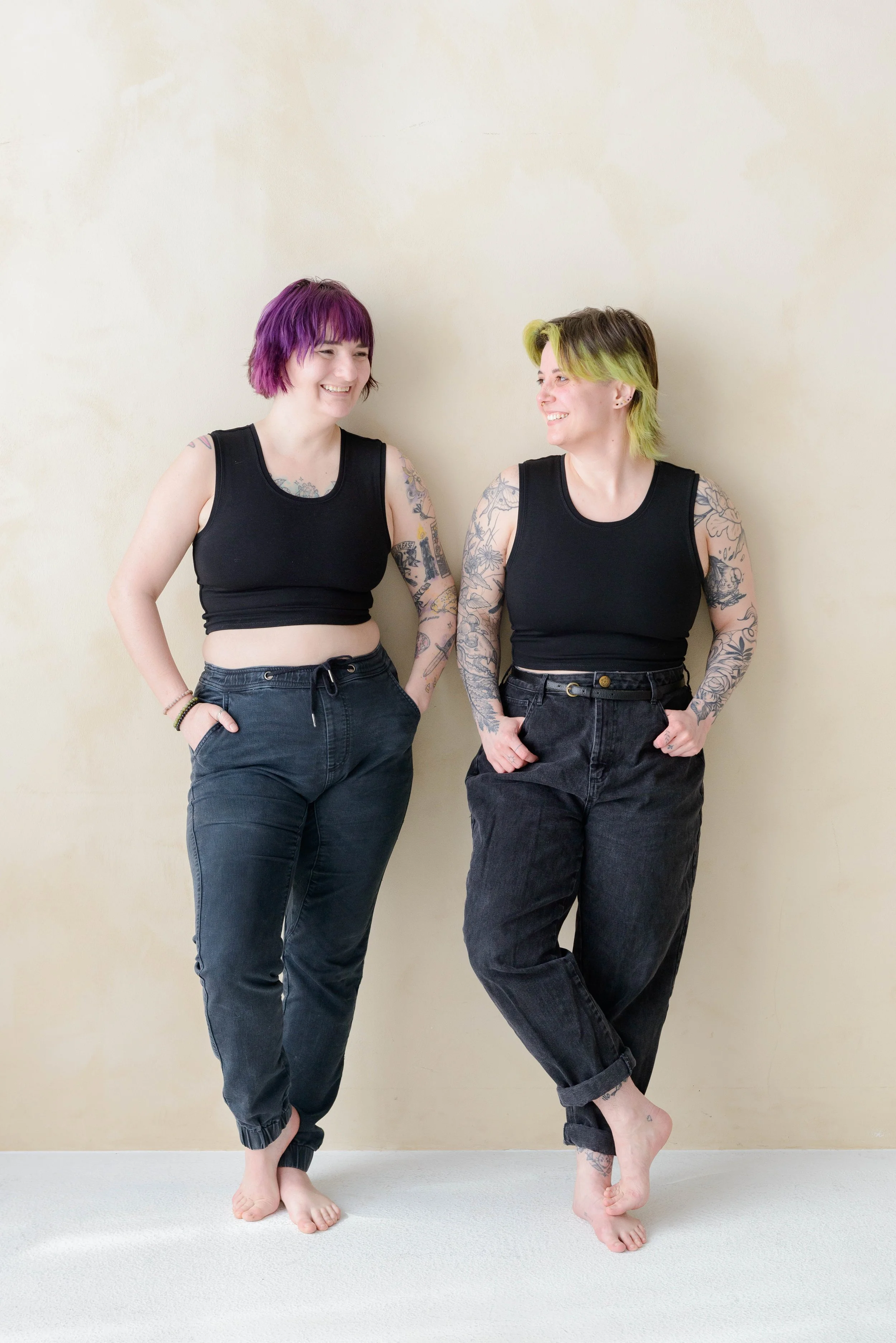 Two nonbinary people with colorful hair and tattoos wearing black chest binders and black pants, standing barefoot against a plain beige wall, smiling and looking at each other.