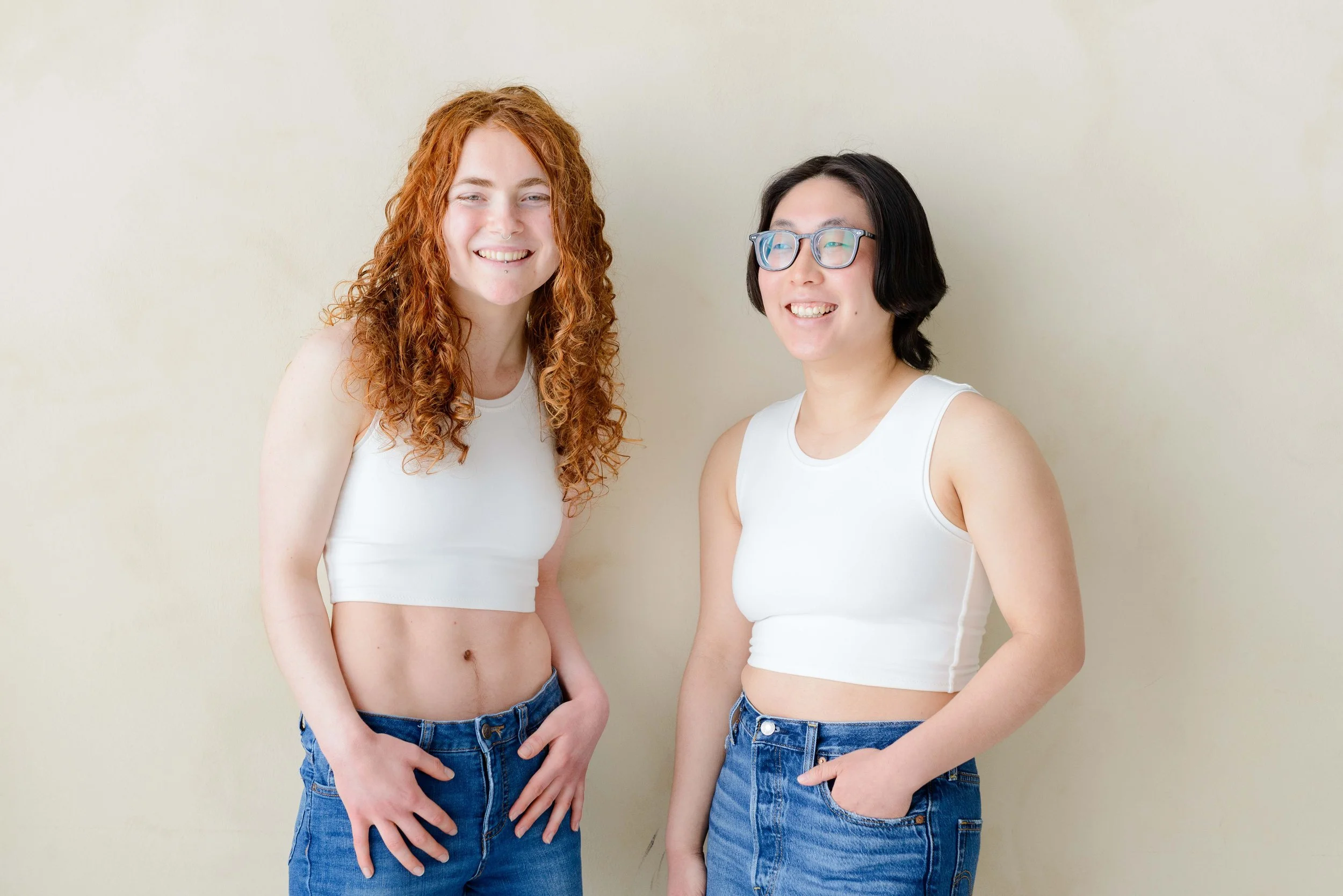 Two nonbinary people standing against a plain beige wall, wearing white chest binders and blue jeans. The person on the left has long curly red hair, a nose ring. The person on the right has short black hair and glasses.