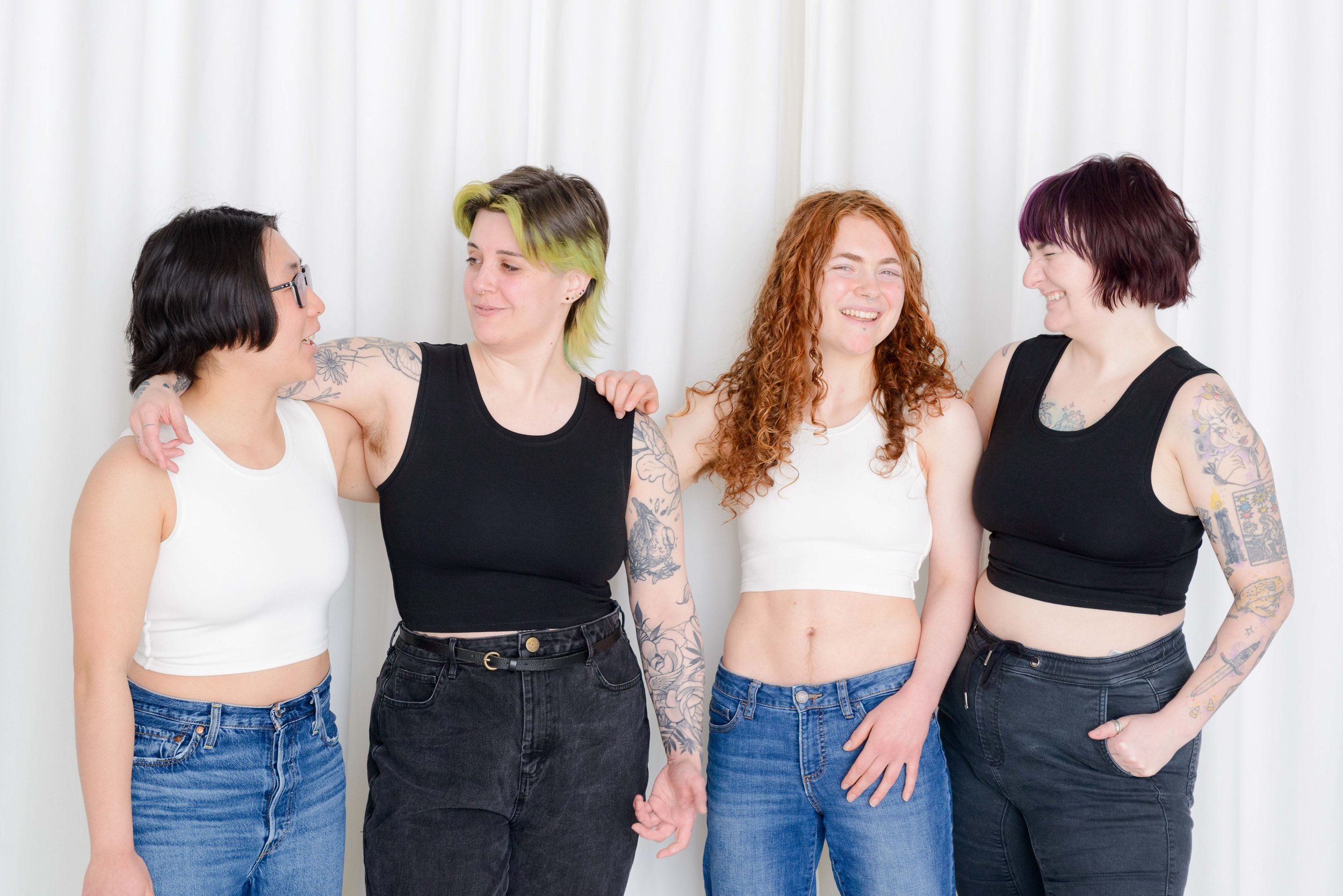Four nonbinary people with tattoos and colorful hair, wearing chest binders and jeans, standing together and smiling, in front of a plain white curtain backdrop. Two are wearing white chest binders and two are wearing black, alternating.