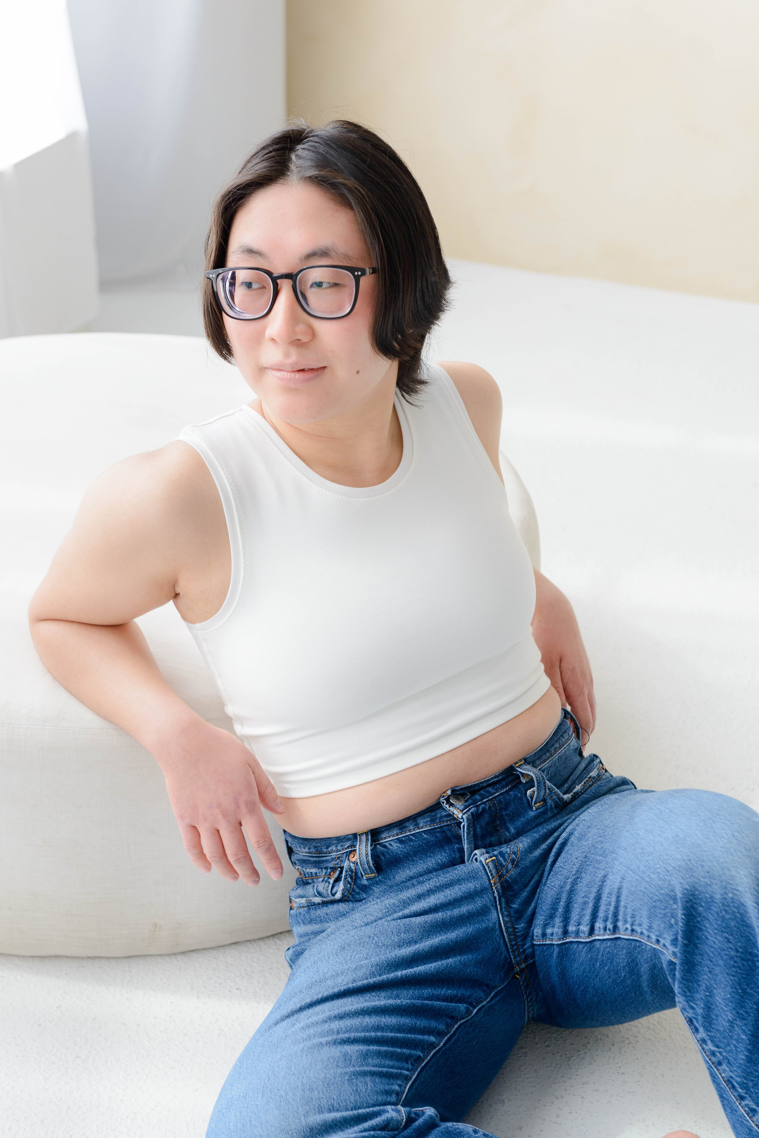A nonbinary person with short dark hair, wearing glasses and a white chest binder, seated, leaning on a round white cushion with their left elbow resting on the seat behind them and their right hand on their lap, in a bright room with natural light.