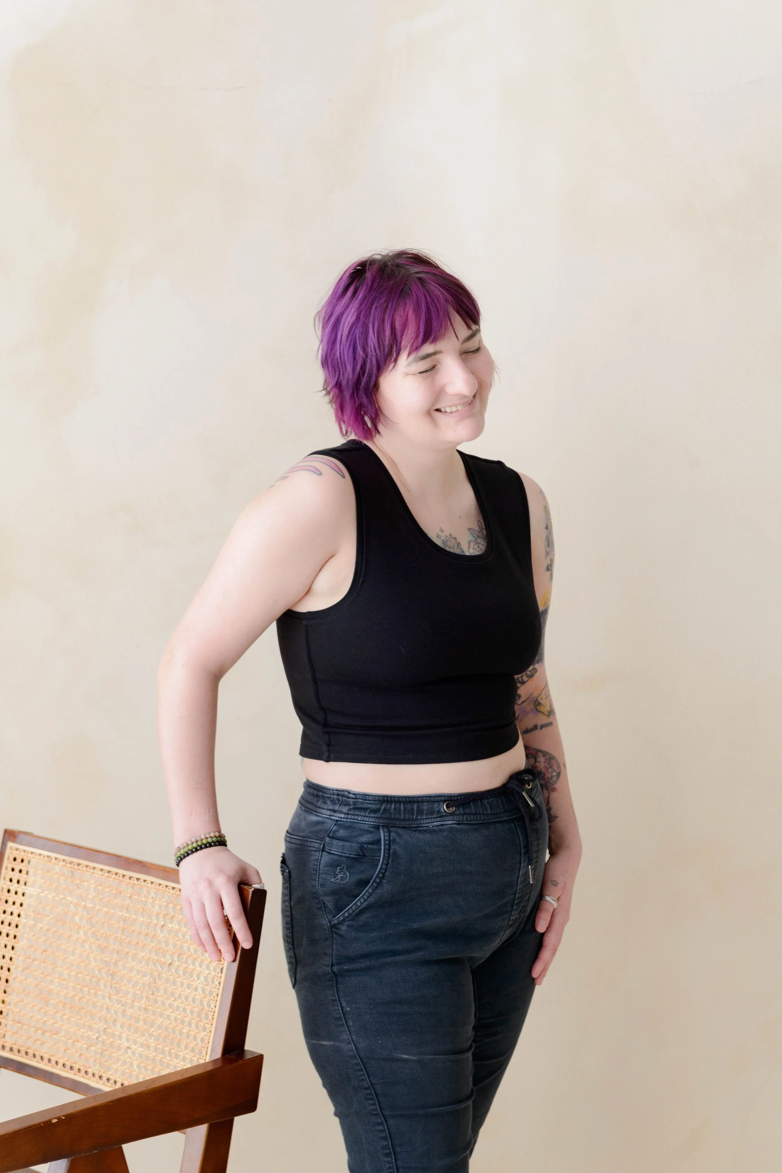 A nonbinary person with short purple hair, wearing a black chest binder and jeans, smiling with their eyes closed, standing next to a wooden chair with a cane backrest, against a beige wall.