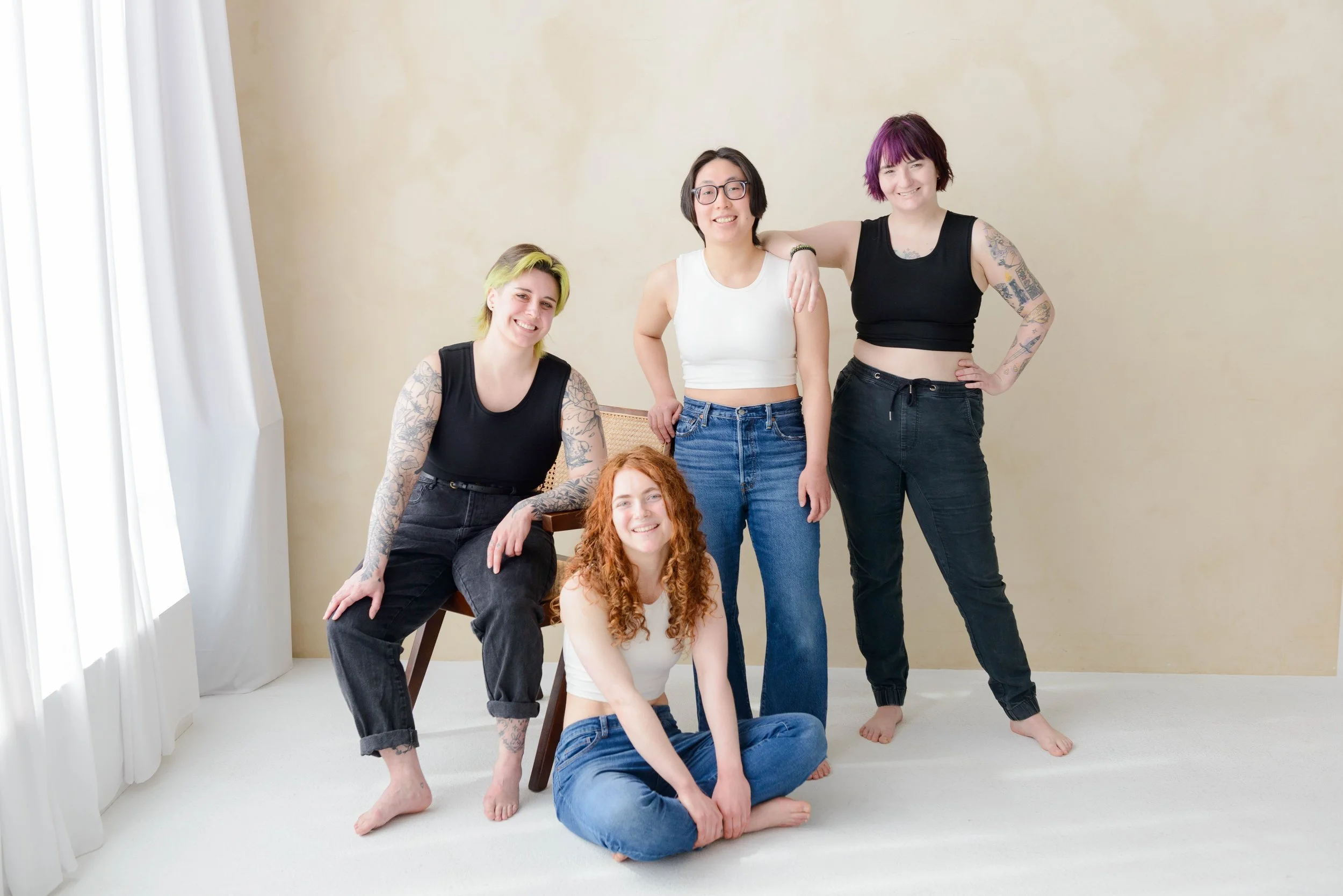 Four models wearing chest binders, two in white and two in black. From left to right, one model is seated on a chair, next is seated cross-legged on the floor, final two are standing.