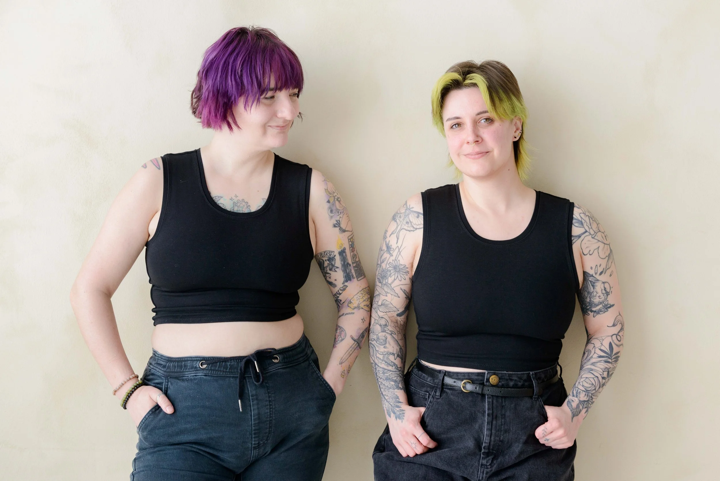 Two nonbinary people with tattoos wearing black chest binders and jeans standing against a beige wall. One has purple hair and the other has greenish hair with brown roots.