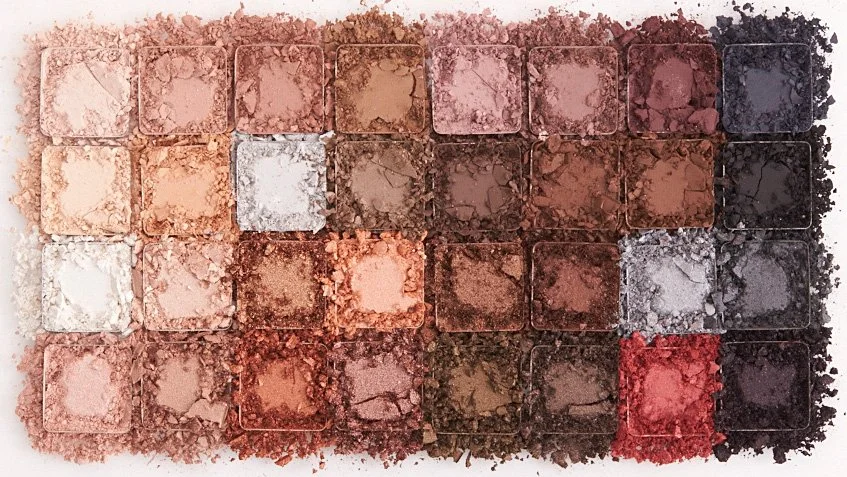 An eyeshadow palette with 25 different shades arranged in a 5x5 grid, ranging from light beige to dark brown, gray, and red tones. Los Angeles beauty product photography.
