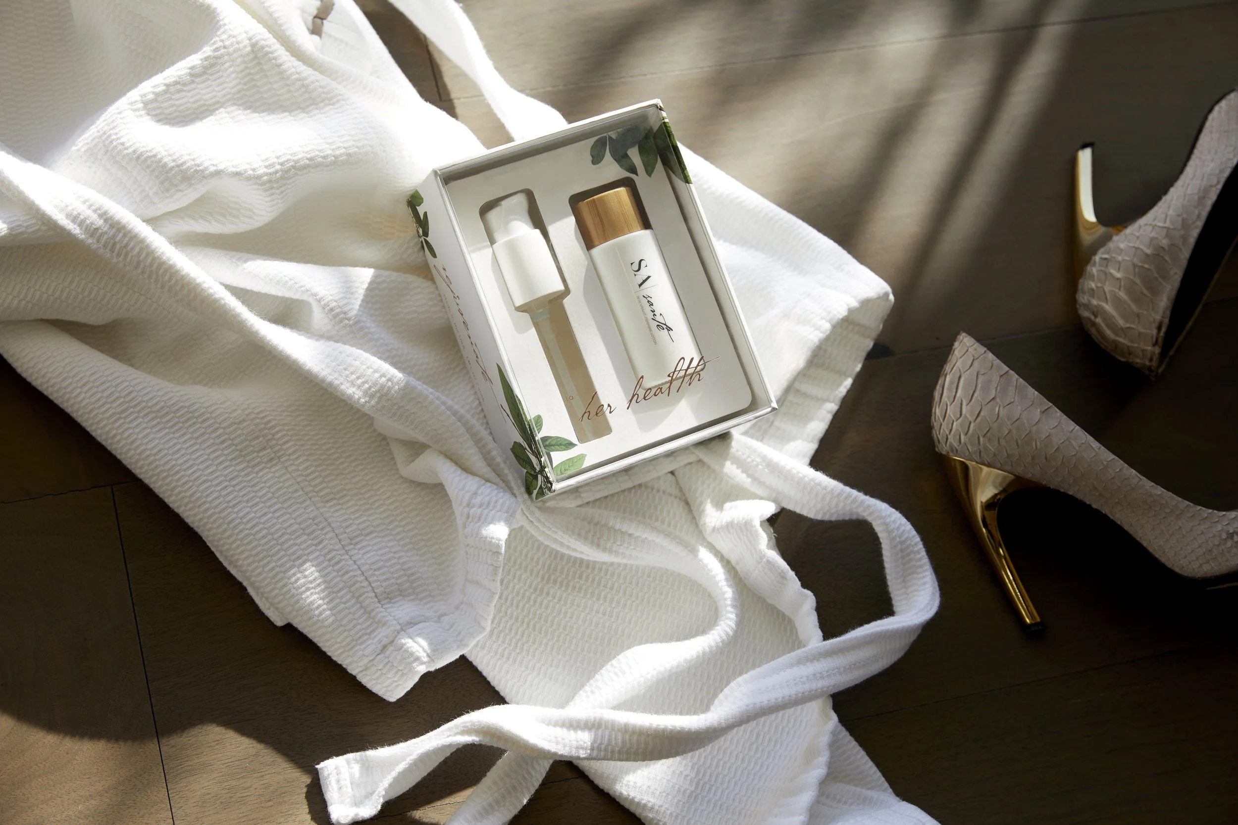 A boxed skincare set with a bottle and roller, resting on a white towel, with a pair of high-heeled shoes nearby on a wooden floor. Los Angeles beauty product photography.