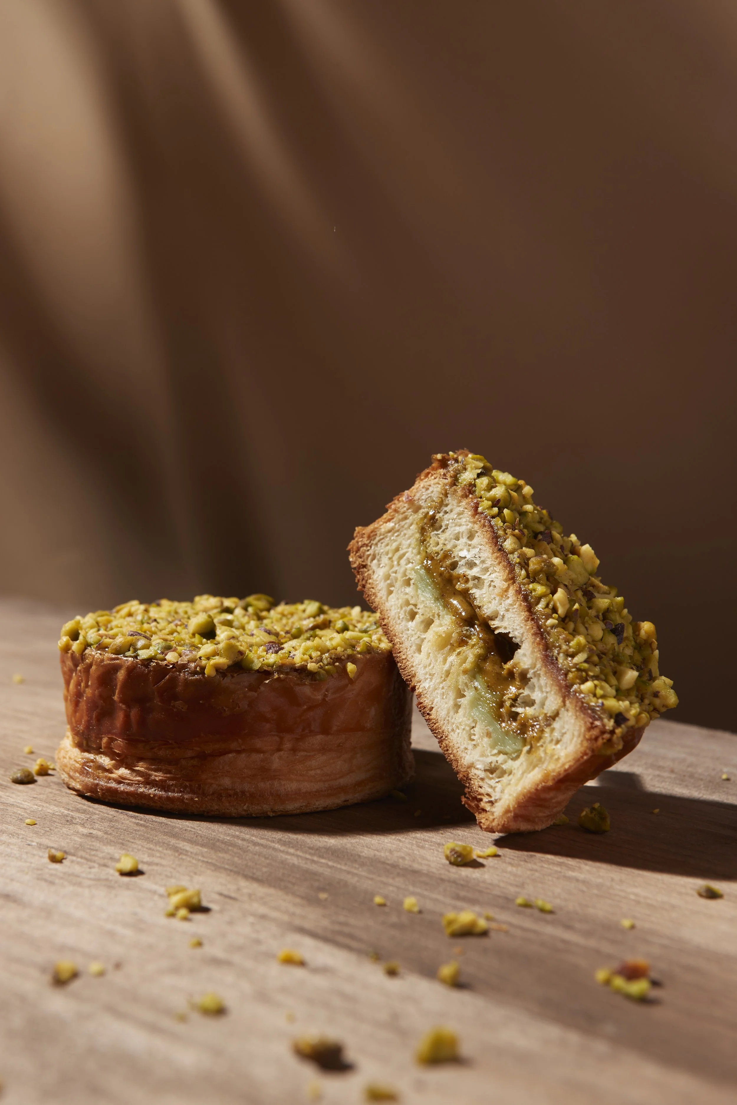 A pistachio pastry from a bakery with a sliced piece revealing a creamy filling, topped with chopped pistachios, on a wooden surface. Los Angeles food photography.