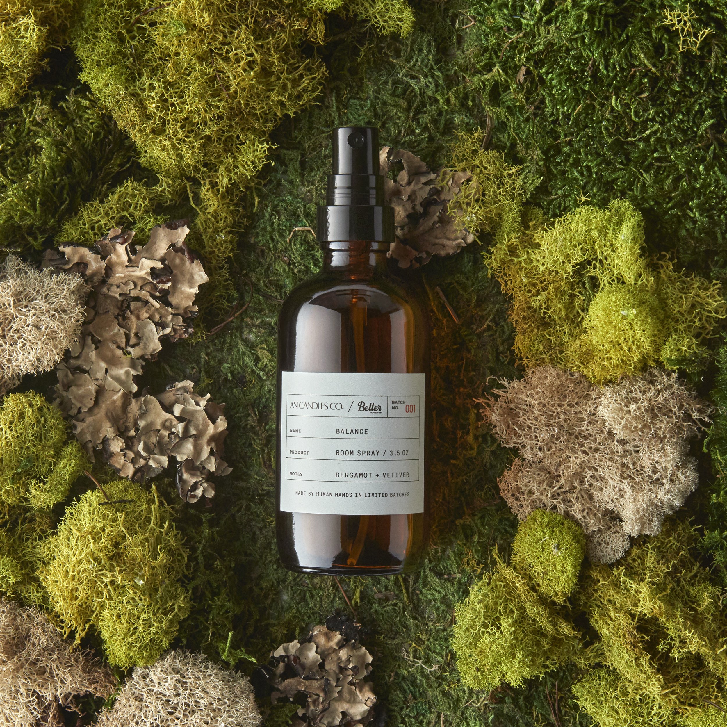A brown spray bottle labeled 'Balance' with notes of bergamot and vetiver, surrounded by green and beige moss and lichen on a forest floor. Los Angeles room spray product photography.