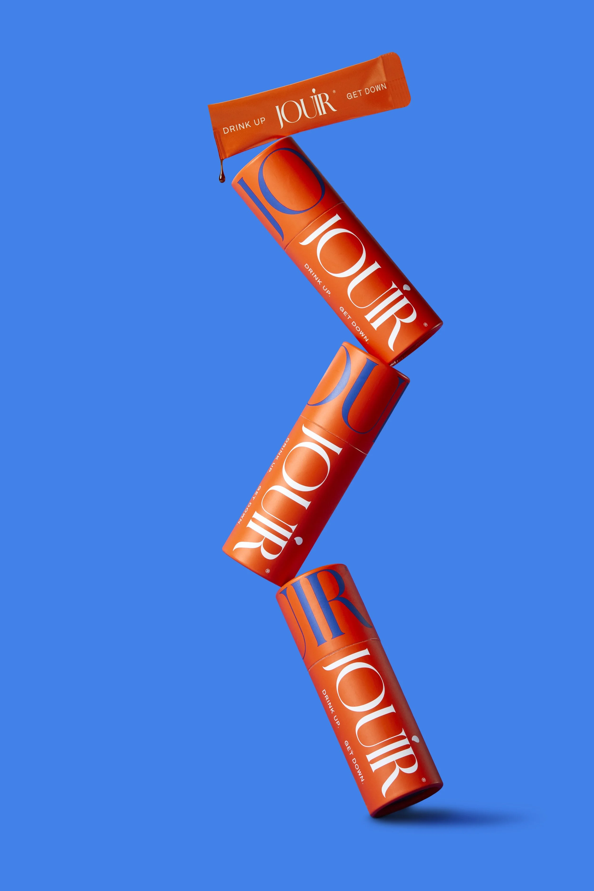 Four orange cylindrical cans with the word 'JOUR' in white and blue text, stacked vertically against a bright blue background, with a small orange stick pack at the top that says 'DRINK UP JOUR GET DOWN'. Los Angeles beauty product photography.