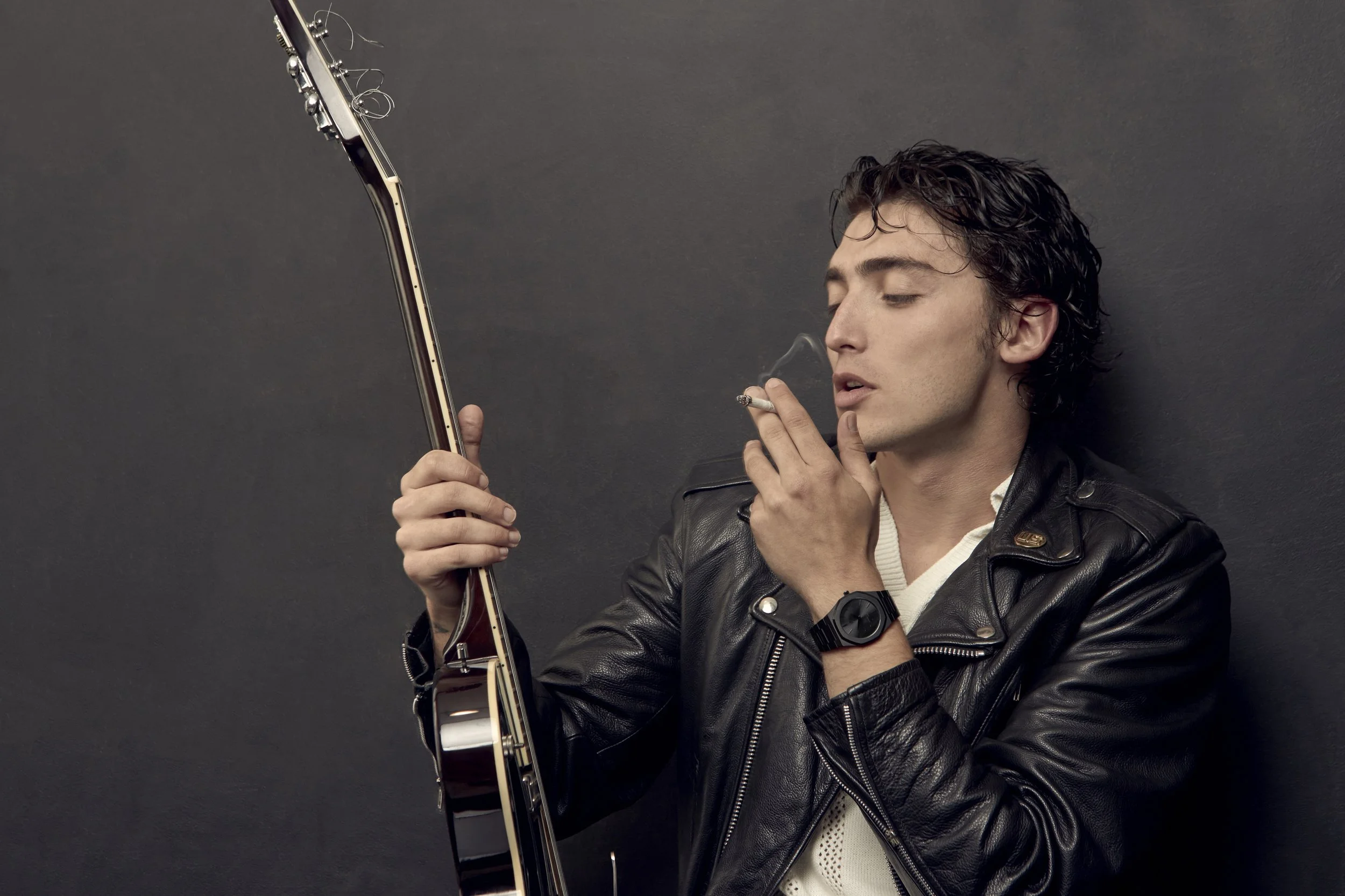A young man in a black leather jacket holding an electric guitar, smoking a cigarette, and standing against a dark gray background.  Los Angeles On-Model fashion photography.