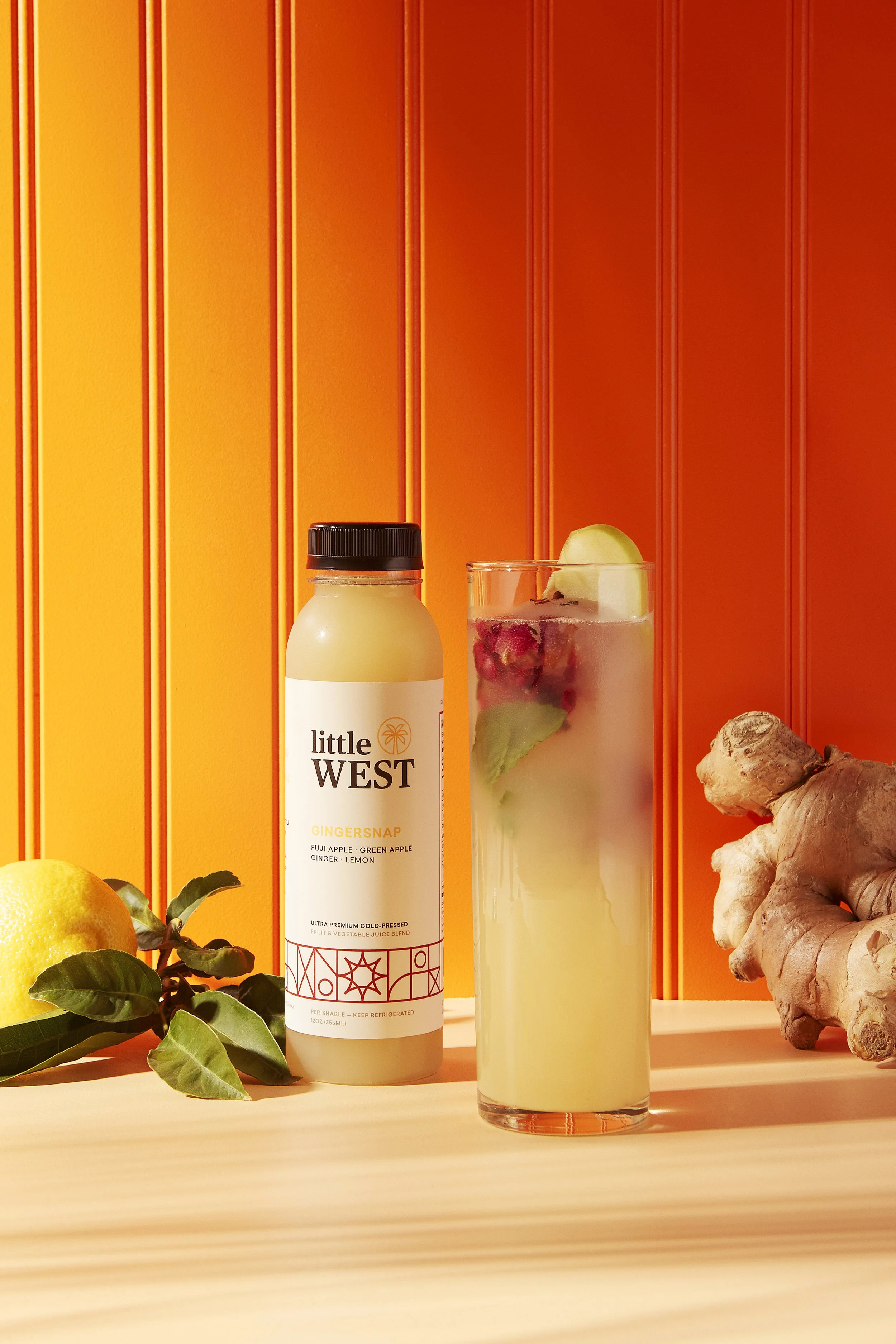 Bottle of Little West Ginger Snap juice, tall glass with a lemon slice, ginger root, lemon, and green leaves on an orange background. Los Angeles beverage and food photography.
