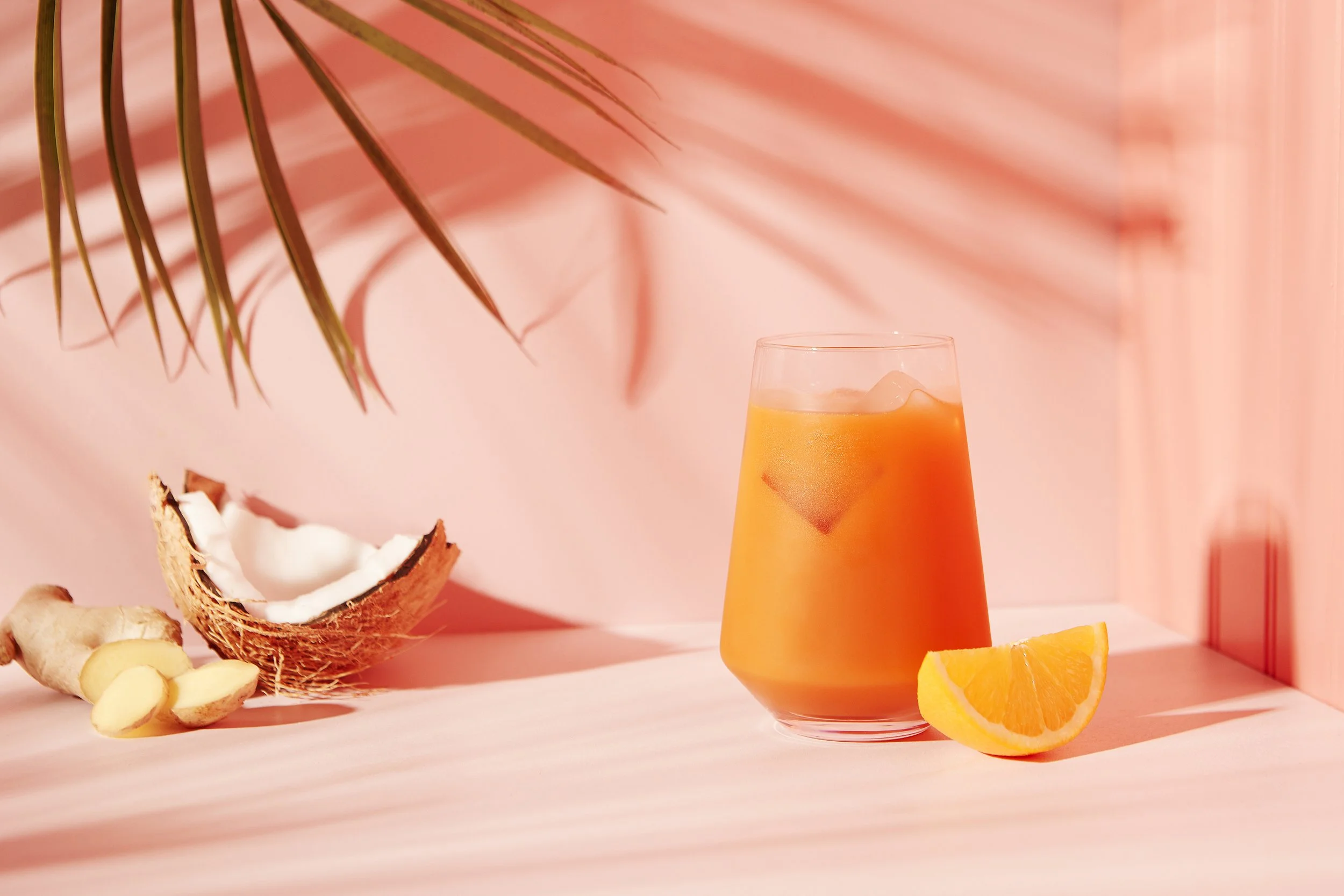 A glass of orange juice with ice cubes, a lemon wedge, a piece of ginger, a coconut half, and a thin palm leaf on a pink background. Los Angeles beverage and food photography.