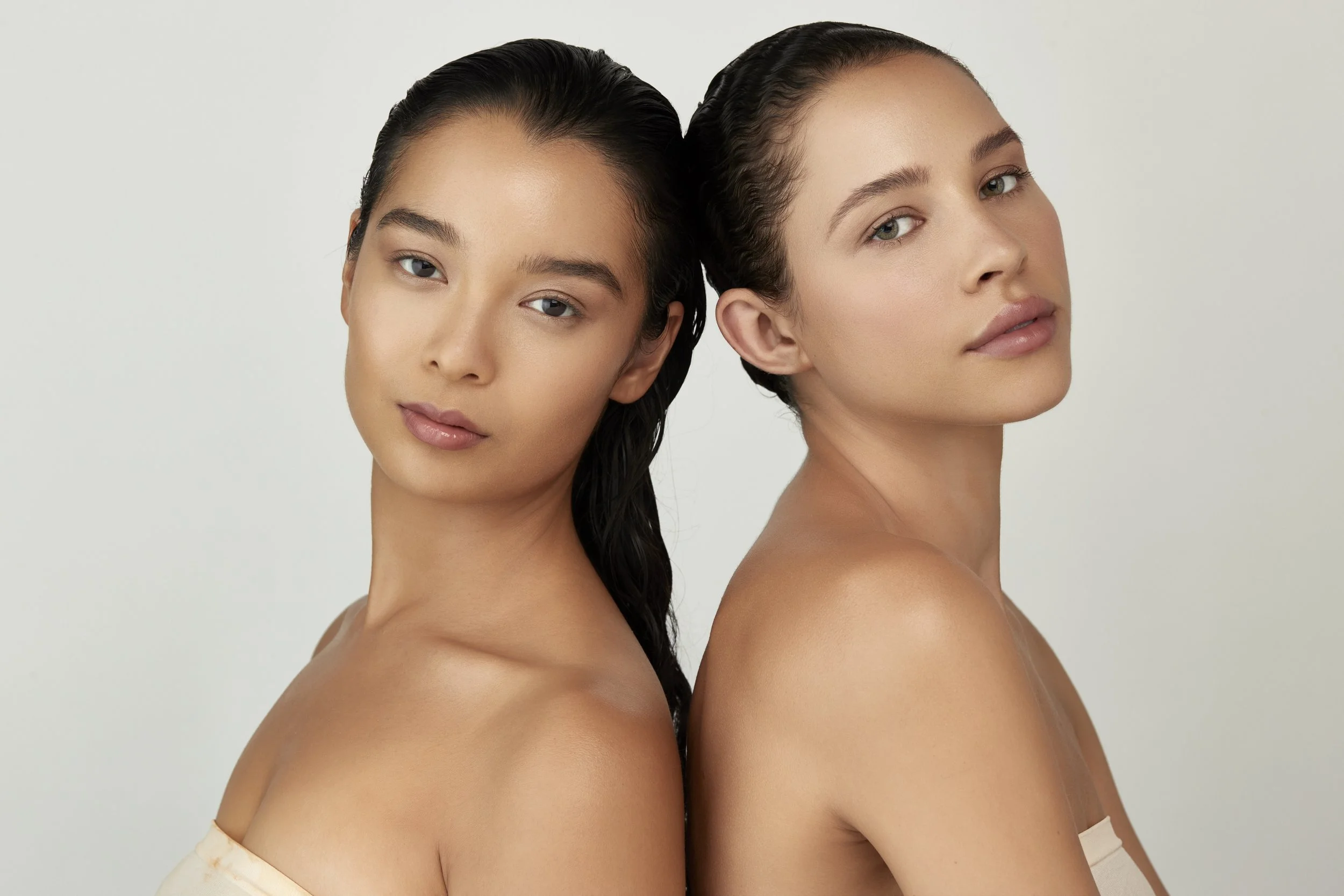 Two young women with wet hair and bare shoulders, facing opposite directions, looking at the camera with neutral expressions. Beaty on-Model Photography Los Angeles.