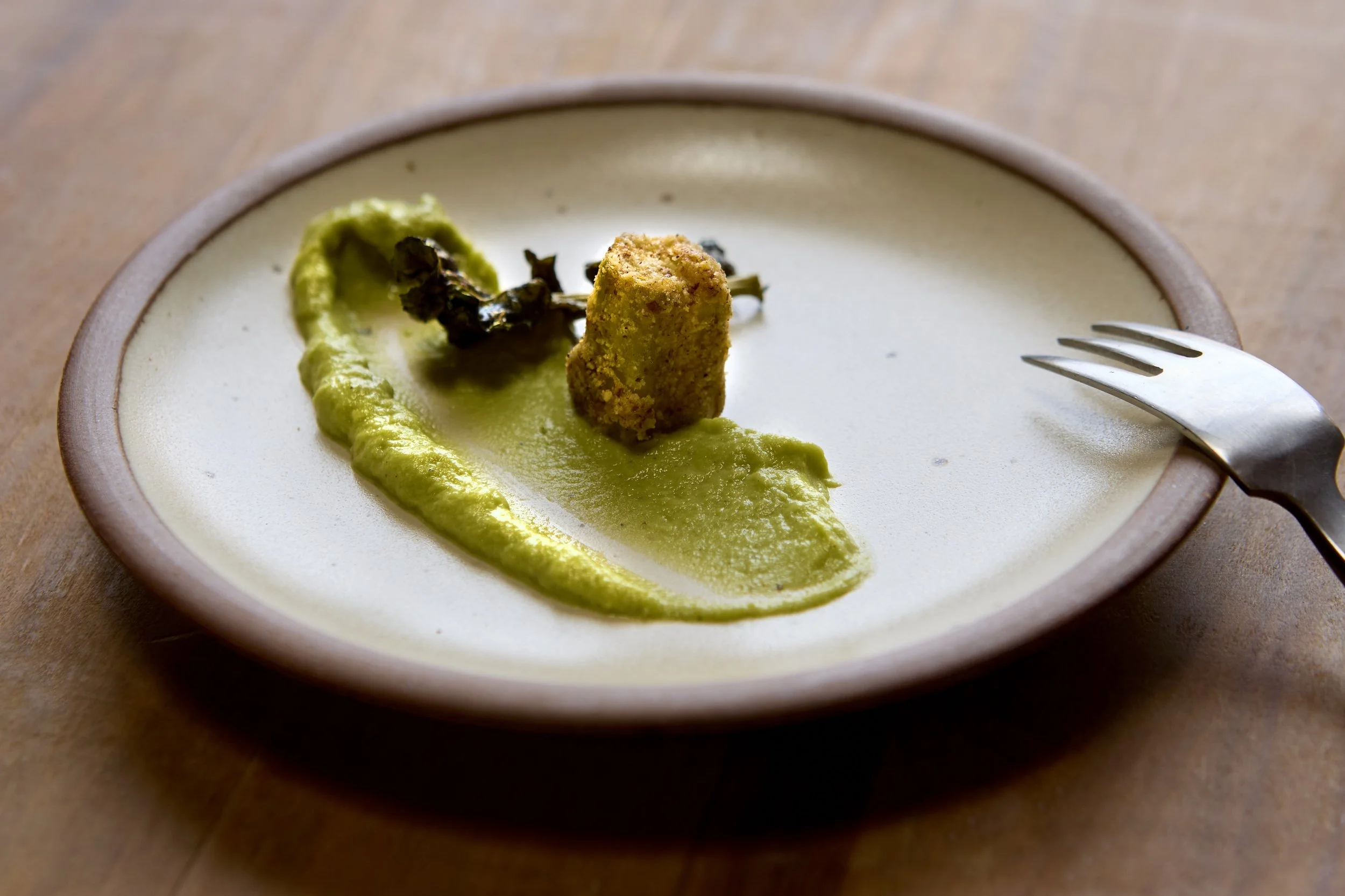 A white plate with a single fried food item, possibly a croquette, on a streak of green sauce, with a small piece of roasted garlic on the sauce. There is a fork on the right side of the plate. Food Photography Los Angeles.