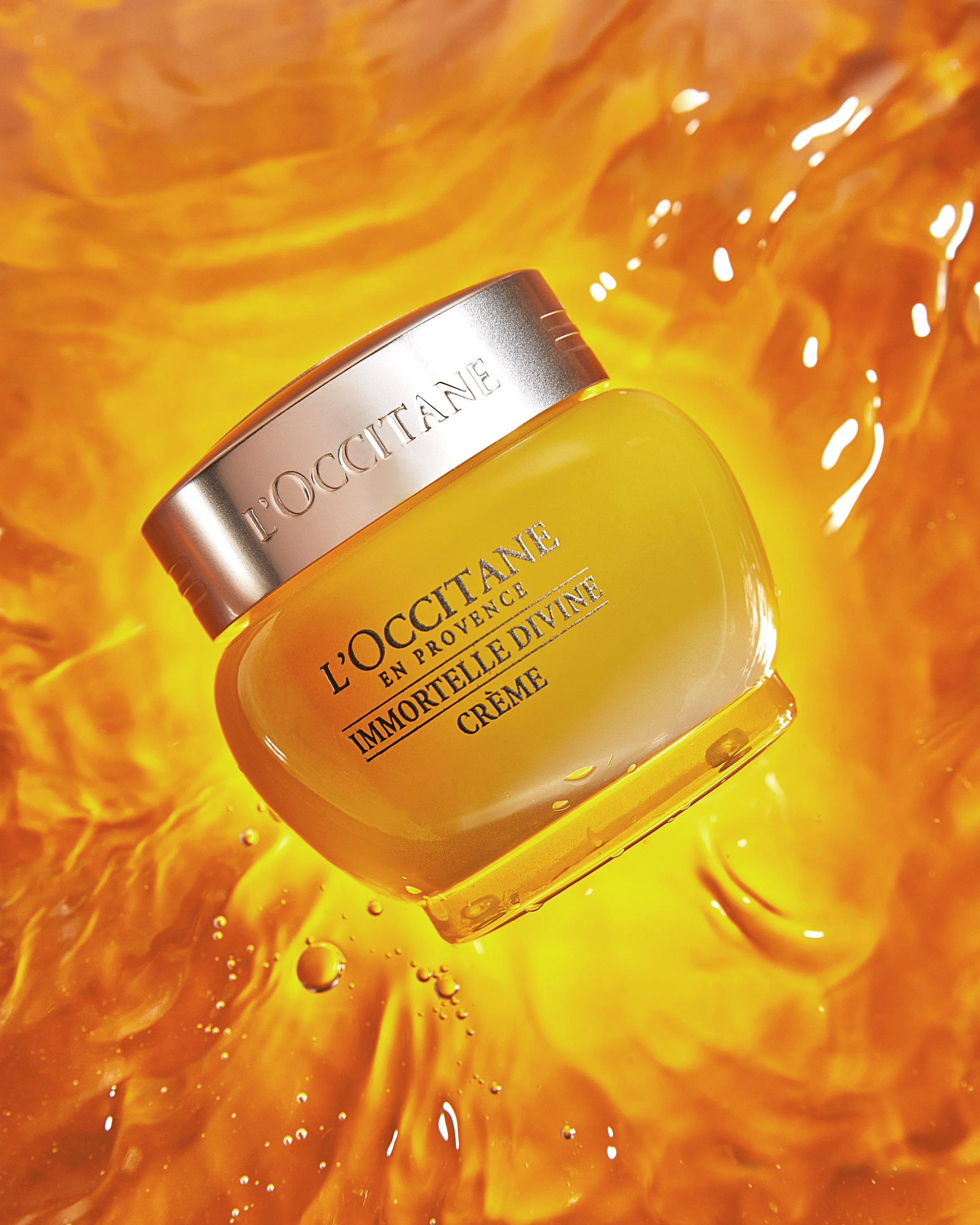 A jar of L'Occitane Immortelle Divine Creme floating in an orange, molten-like background with a glossy surface and bubbles. Beaty Photography Los Angeles.
