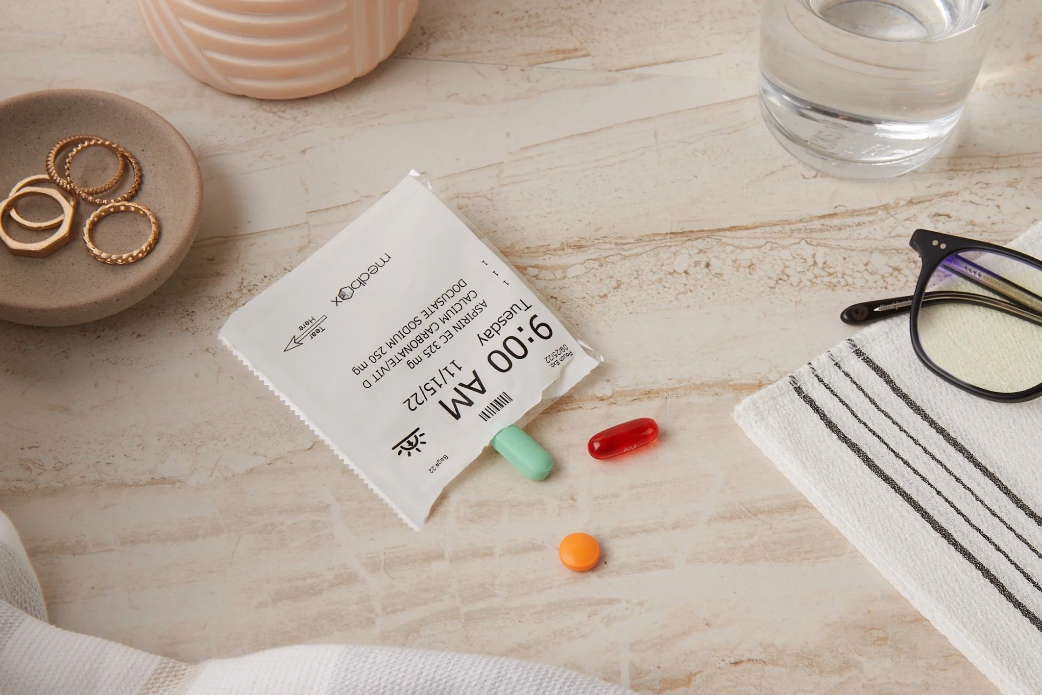 A prescription medication packet with two pills, a glass of water, a pair of eyeglasses, and some jewelry on a light-colored surface. Los Angeles pharma product photography.