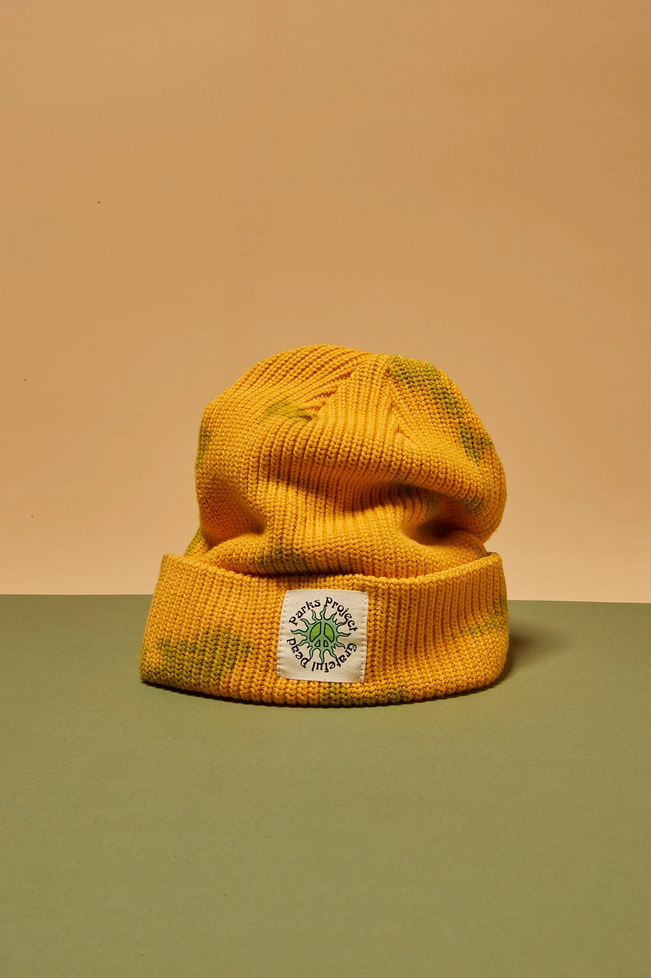 Yellow knitted beanie hat with a Greenpeace logo patch, placed on a green surface against a light pink and beige background. Fashion flatlay photography Los Angeles.