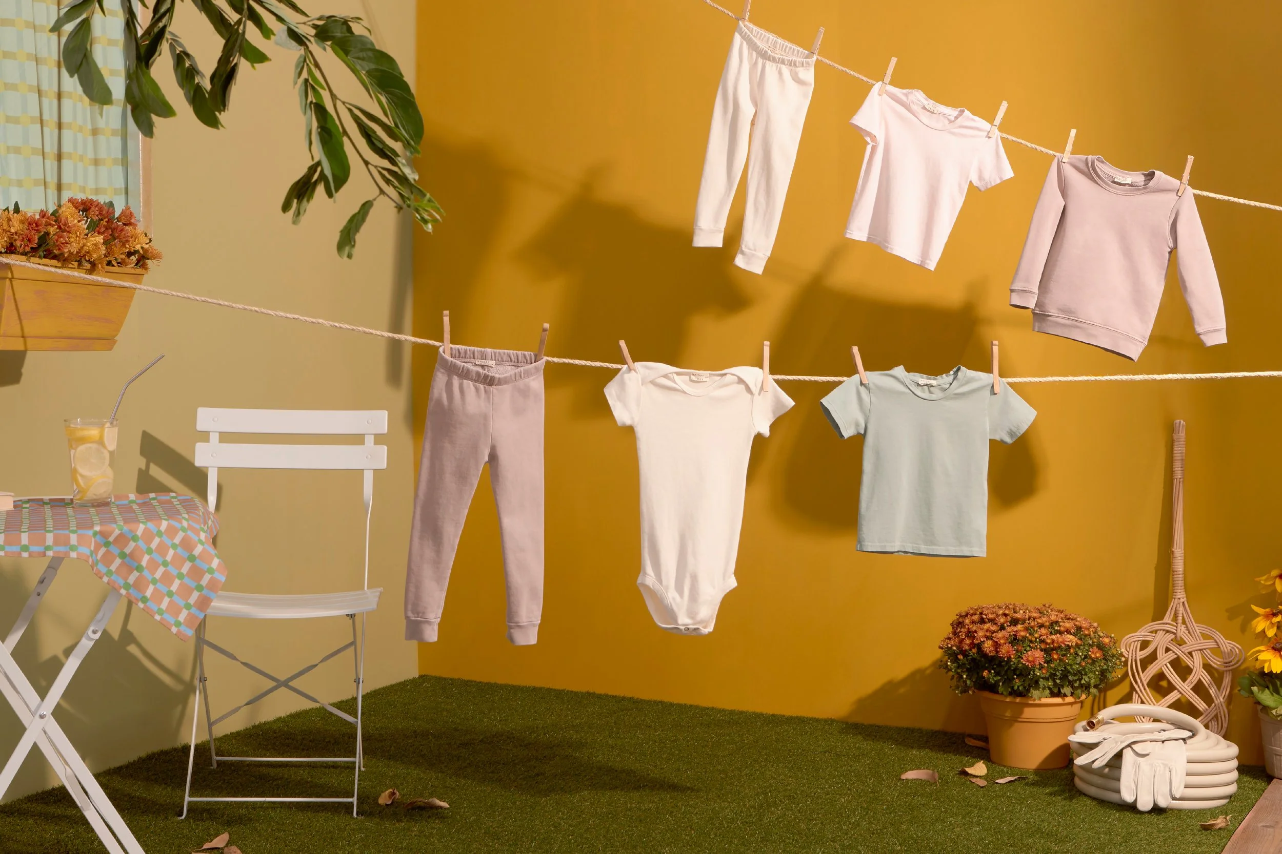 Clothes hanging on a clothesline outdoors with a yellow wall and gardening items nearby. Baby clothes fashion flatlay photography Los Angeles.