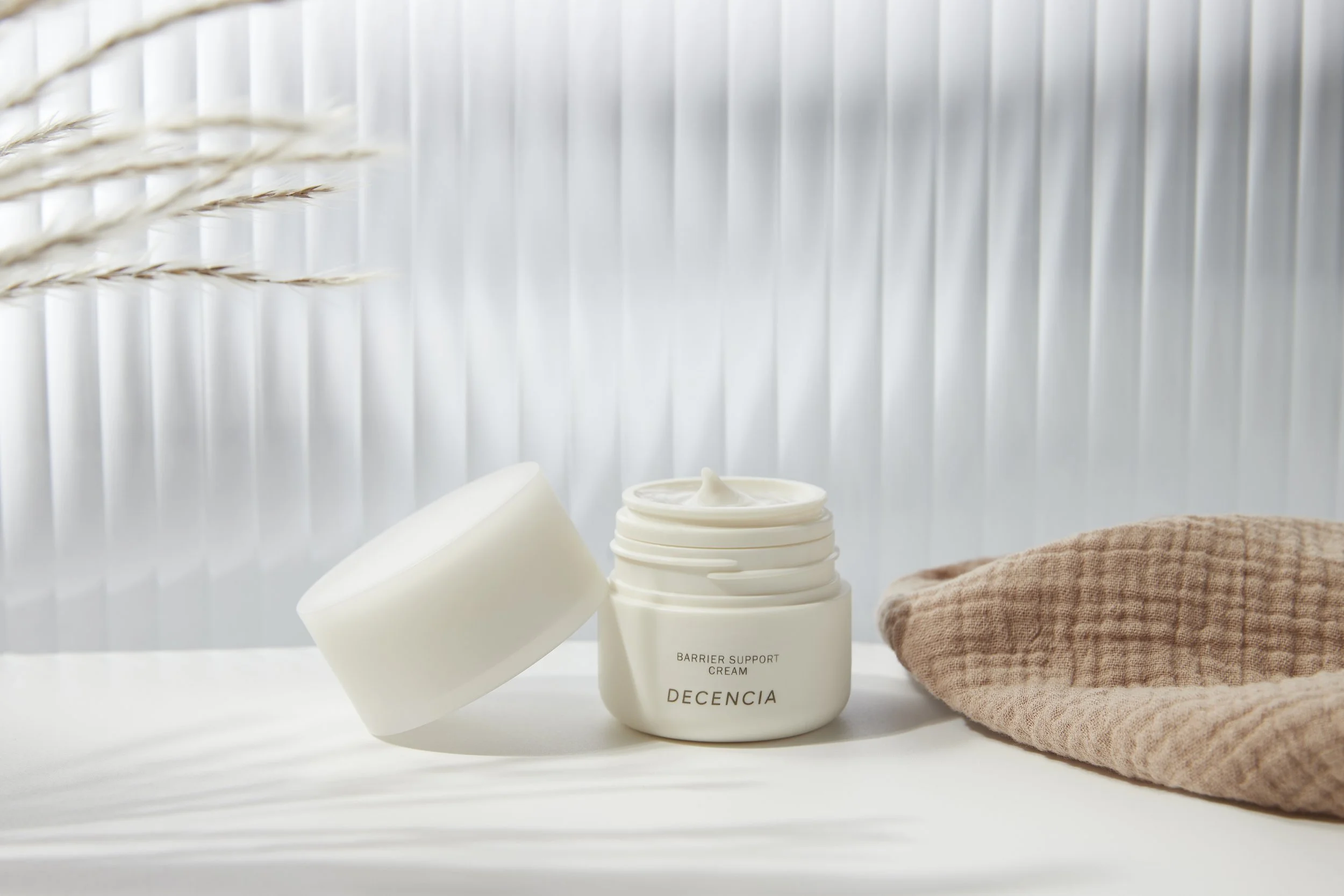 Open jar of Decencia Barrier Support Cream next to the jar's lid and a pink textured cloth on a white surface with a ribbed white background. Beaty Product Photography Los Angeles.