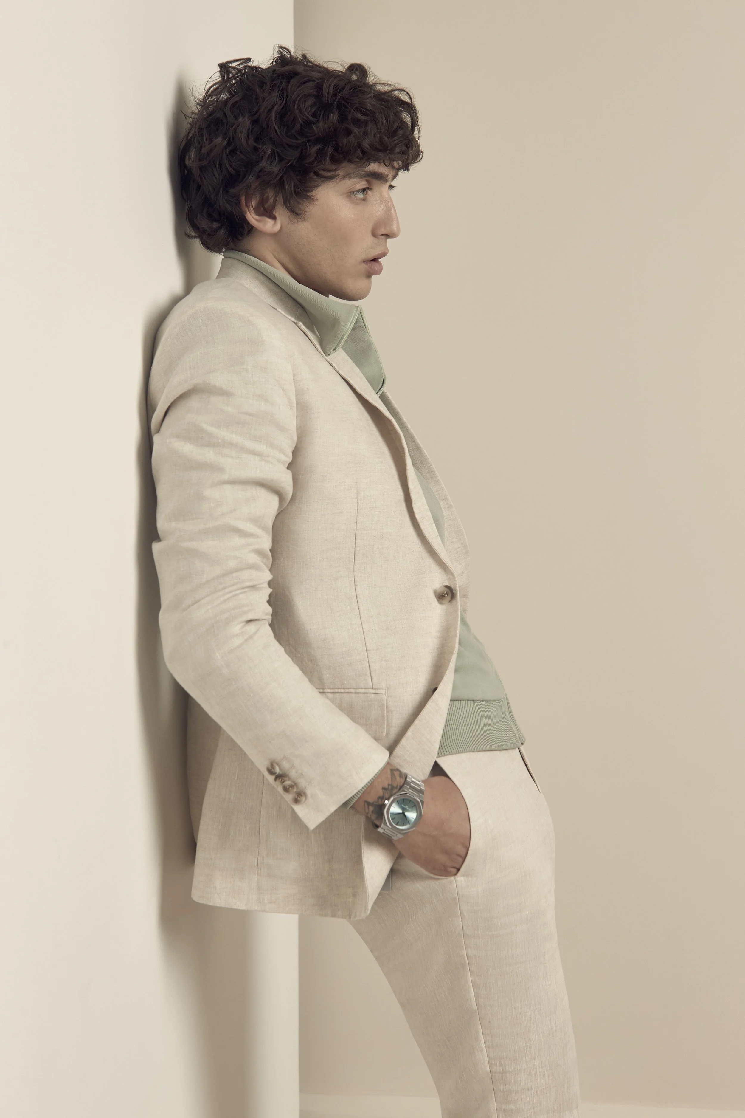 A young man with curly hair leaning against a wall, dressed in a beige suit with a light green shirt and watch, looking to the side. Los Angeles On-Model fashion photography.