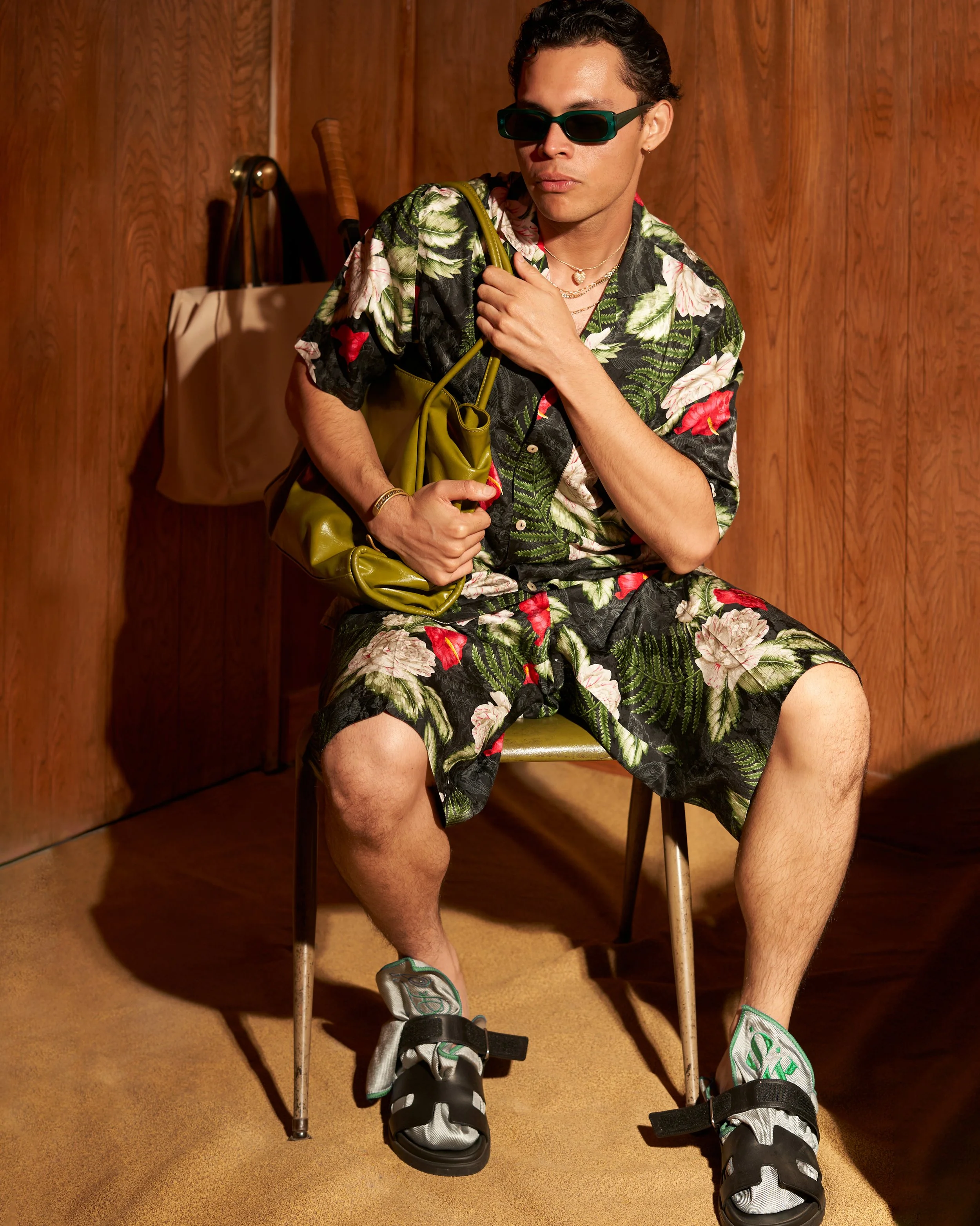 A young man sitting on a chair wearing a floral Hawaiian shirt and matching shorts, black sandals with socks, dark sunglasses, holding a green purse, in a room with wood-paneled walls. On model fashion photography, Los Angeles.