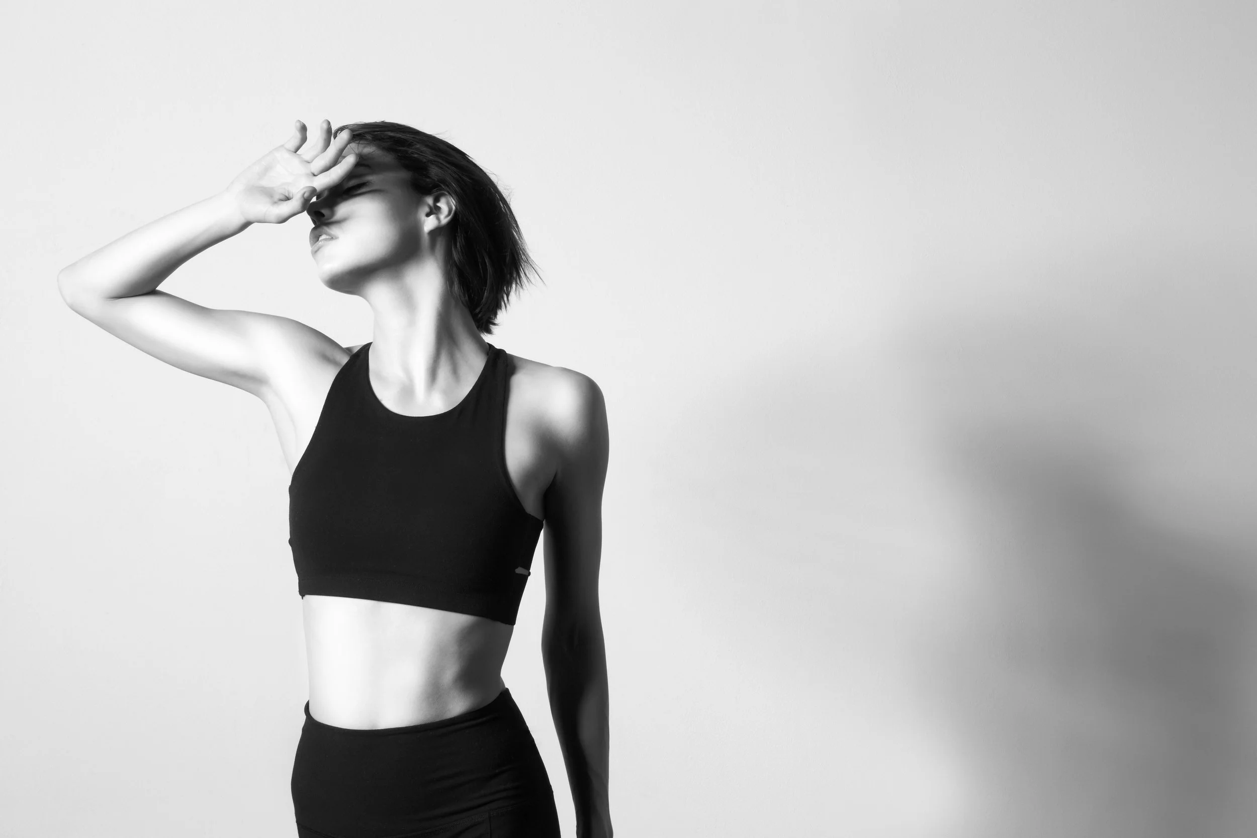 Black and white photo of a woman in athletic wear, with eyes closed, holding her hand to her forehead, and her head tilted back against a plain wall.  Los Angeles On-Model fashion photography.