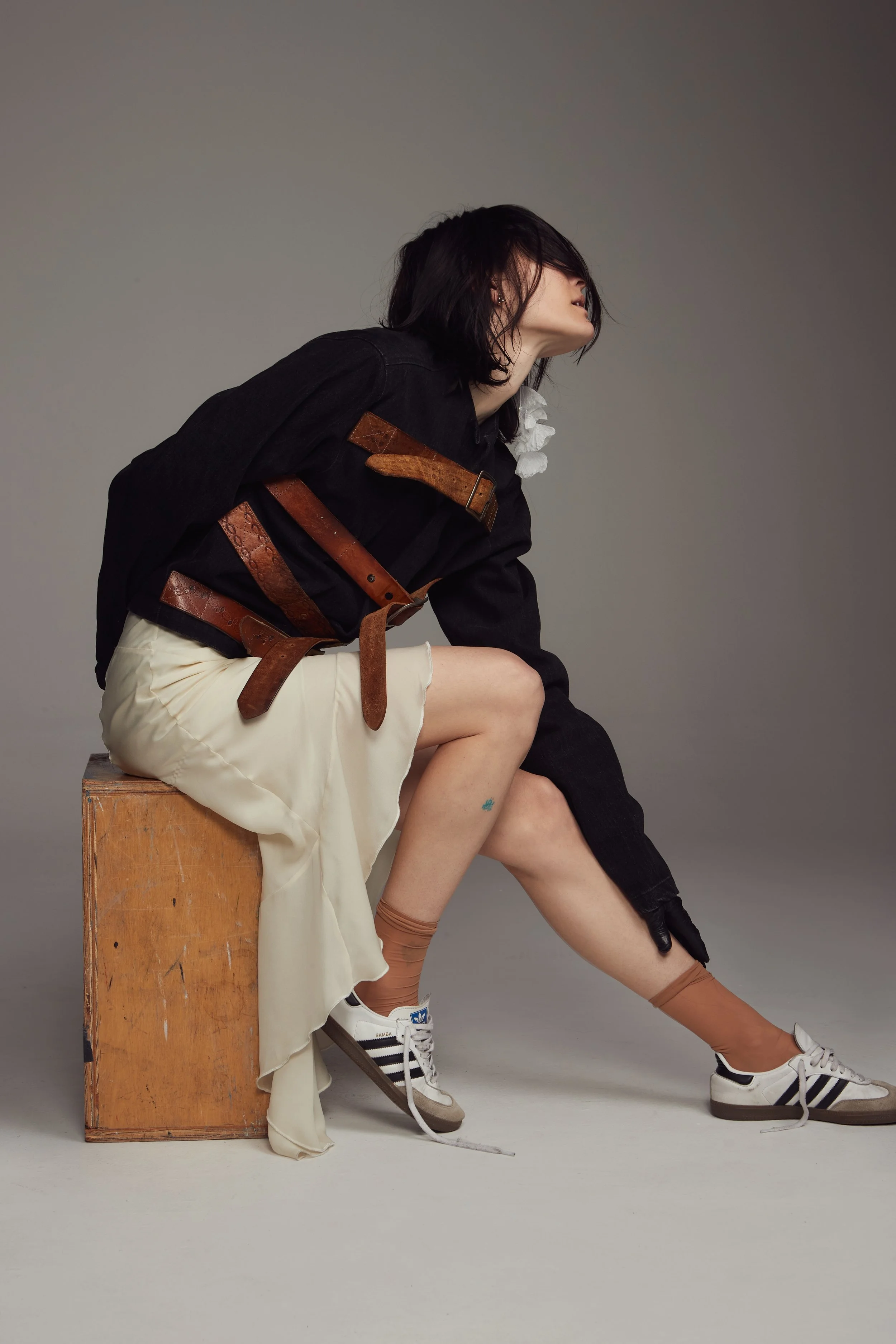 A woman with black hair sits on a wooden box against a plain gray background. She is dressed in a black top with brown belts, a cream-colored skirt, and white sneakers with black stripes.  Los Angeles On-Model fashion photography.