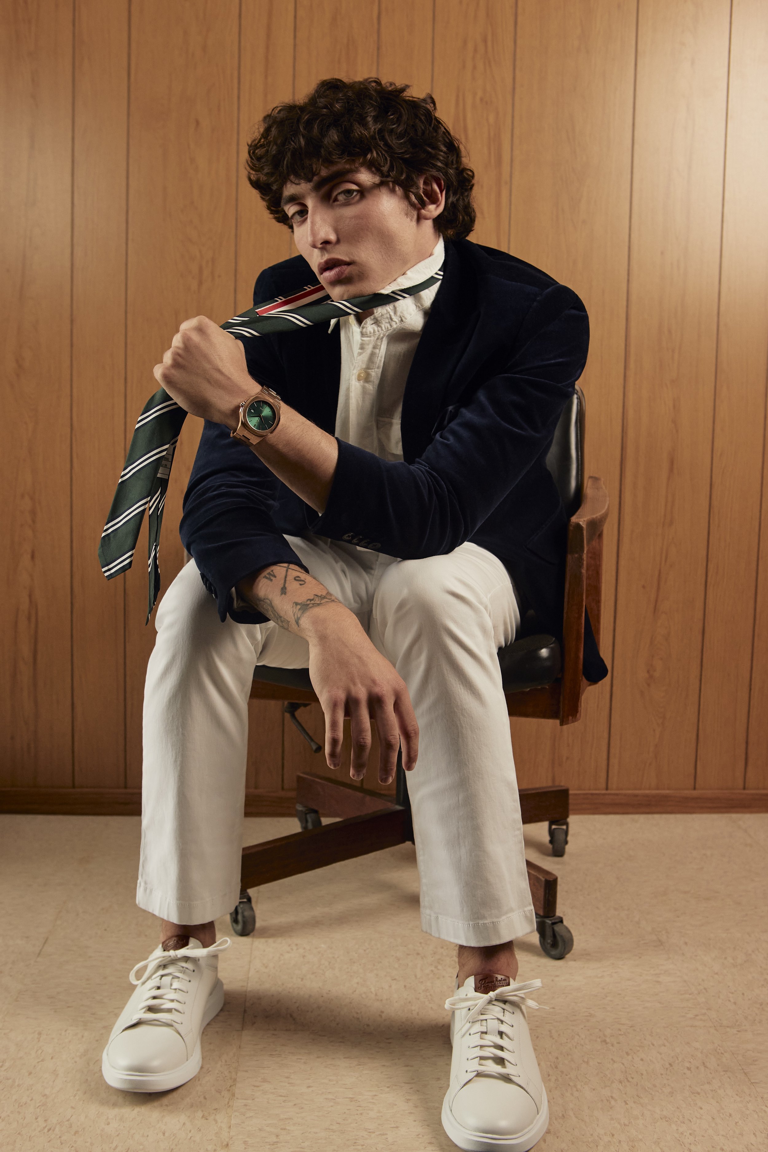 A young man with curly hair sits on a wooden chair in front of a wood-paneled wall. He is wearing a dark blazer, white shirt, beige pants, and white sneakers. On model fashion photography, Los Angeles.