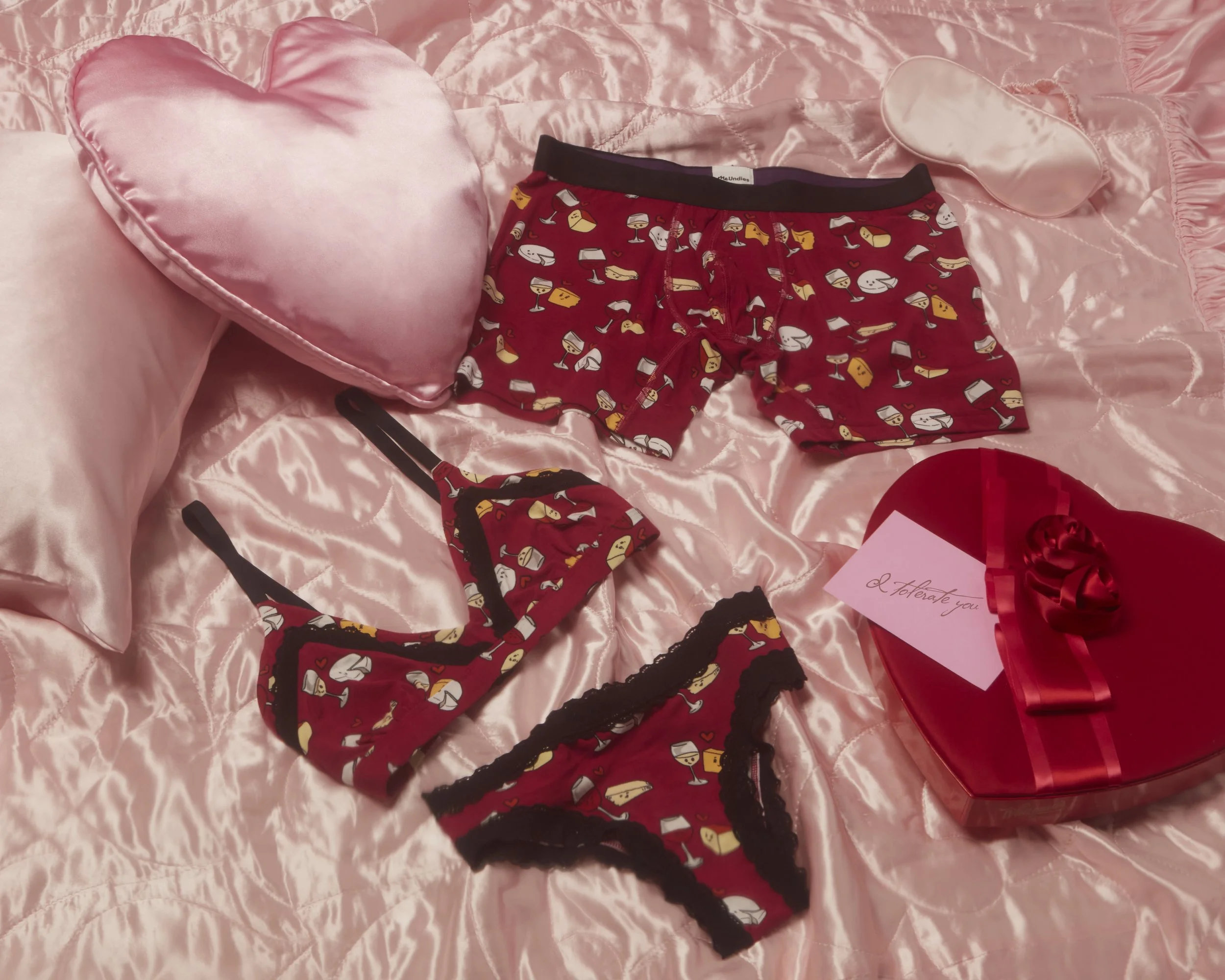 A Valentine's Day gift set on a pink satin bedspread, including red patterned underwear and bra, a pink heart-shaped box with a red ribbon and a note that reads 'I tolerate you.' MeUndies fashion flatlay photography Los Angeles.
