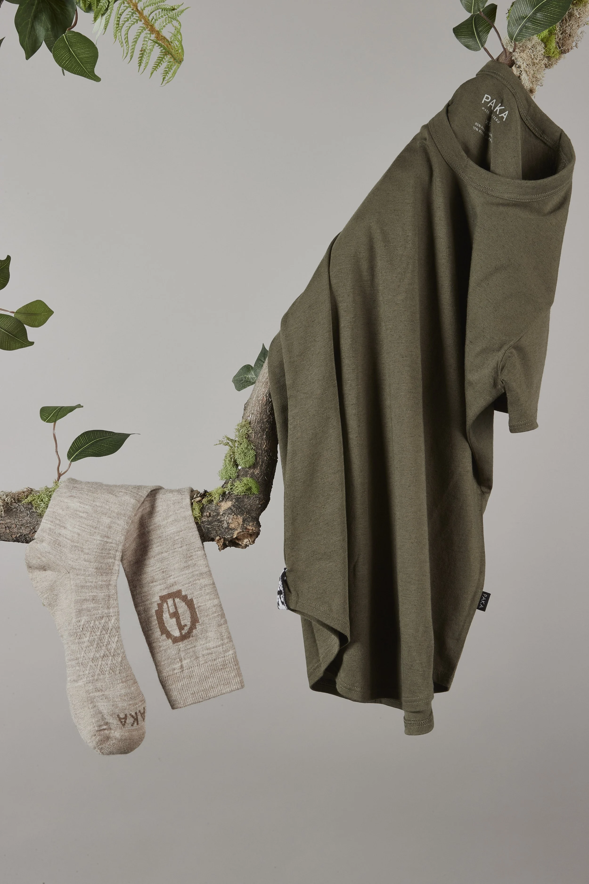 A green PAKA T-shirt hangs on a tree branch with green leaves and moss, alongside a beige sock with brown logo and text, all set against a plain gray background. Fashion flatlay photography Los Angeles.
