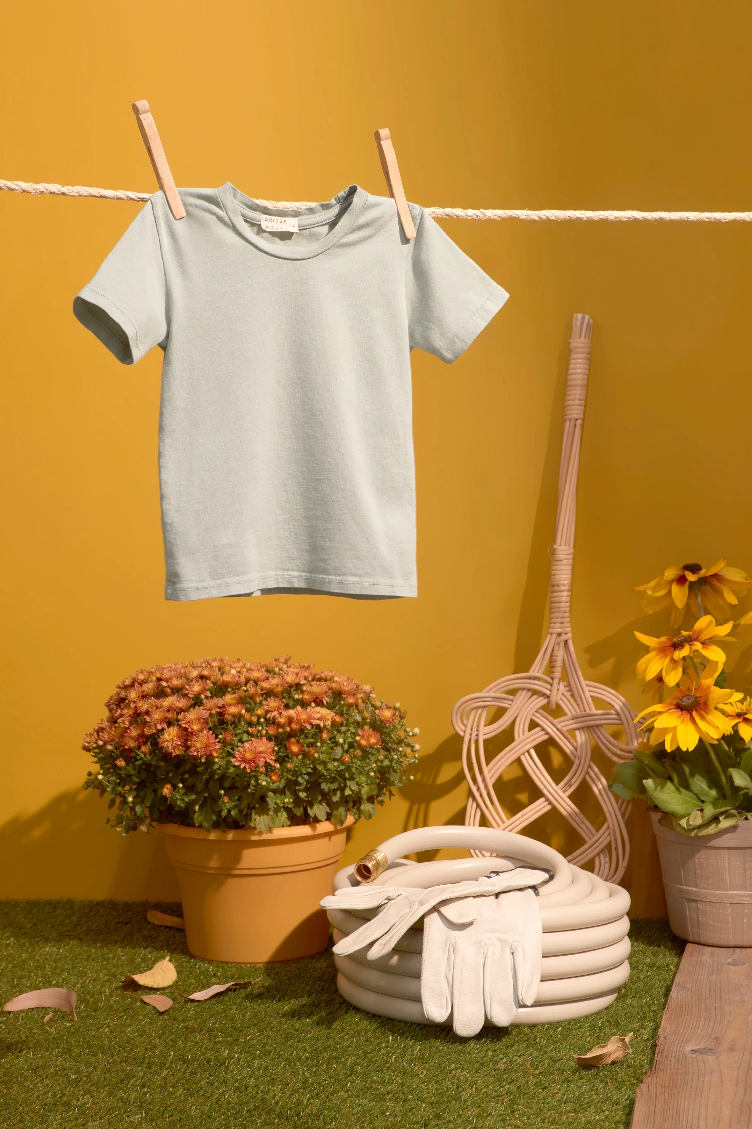 An indoor display with a white T-shirt hanging on a clothesline, surrounded by potted flowers, gardening tools, and a garden hose, against a yellow wall. Product photography Los Angeles.