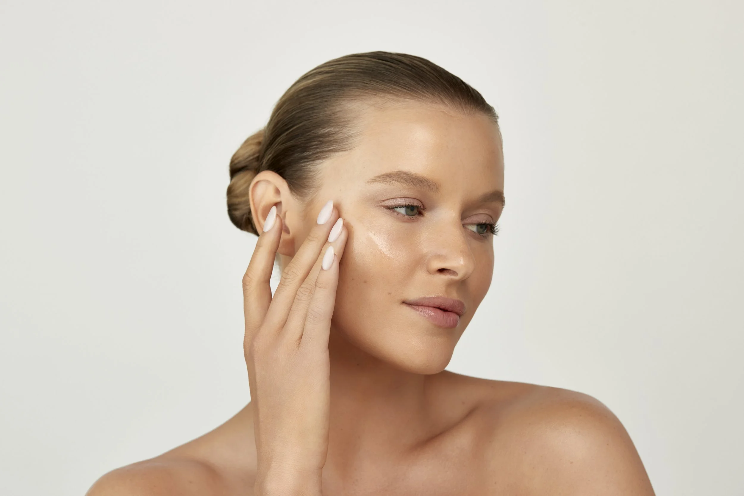 A woman with light skin and brown hair elegantly pulled back, applying skin cream to her face. Los Angeles beauty product photography.