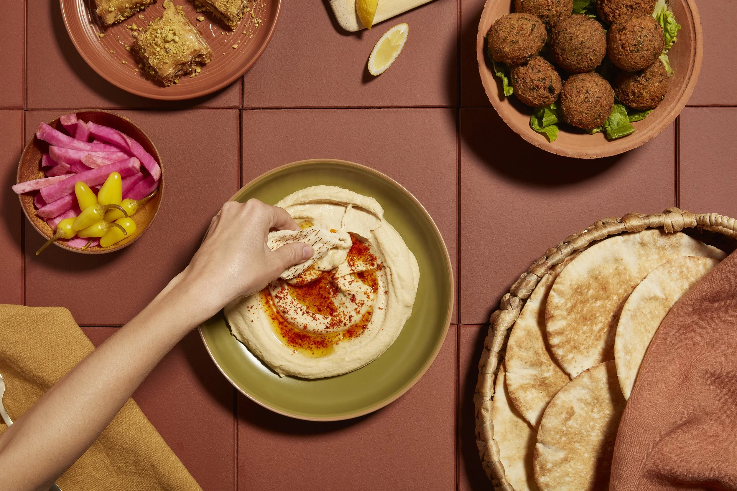 A variety of Mediterranean foods, including pita bread, hummus with paprika, falafel balls on a bed of lettuce, pickled pink turnips with yellow peppers, fried pastries, and lemon wedges, are arranged on a terracotta-colored table food photography.