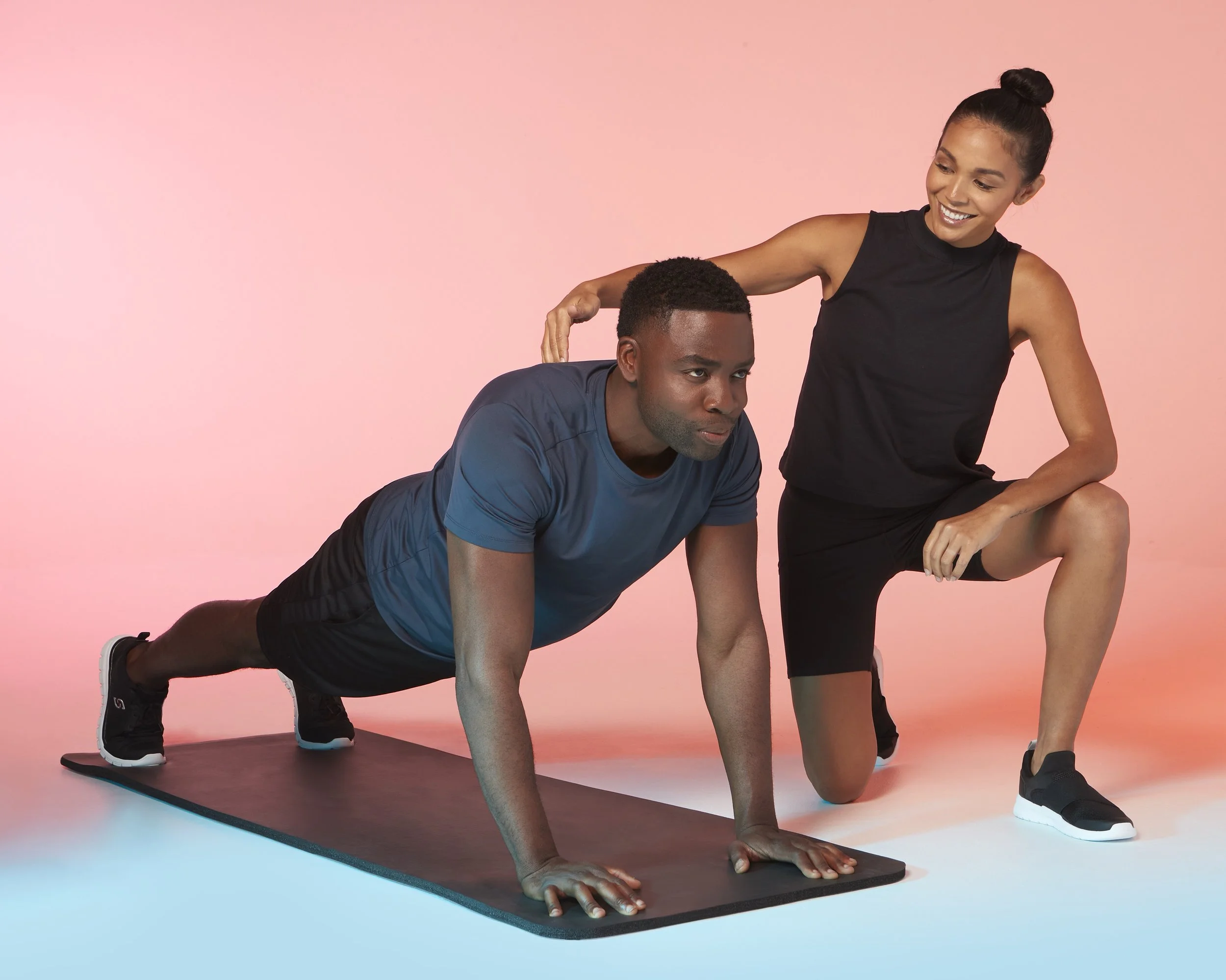 A man in a blue T-shirt and black shorts doing a plank exercise on a yoga mat, guided by a woman in a black sports top and shorts, on a pink background.  Los Angeles On-Model fitness photography.