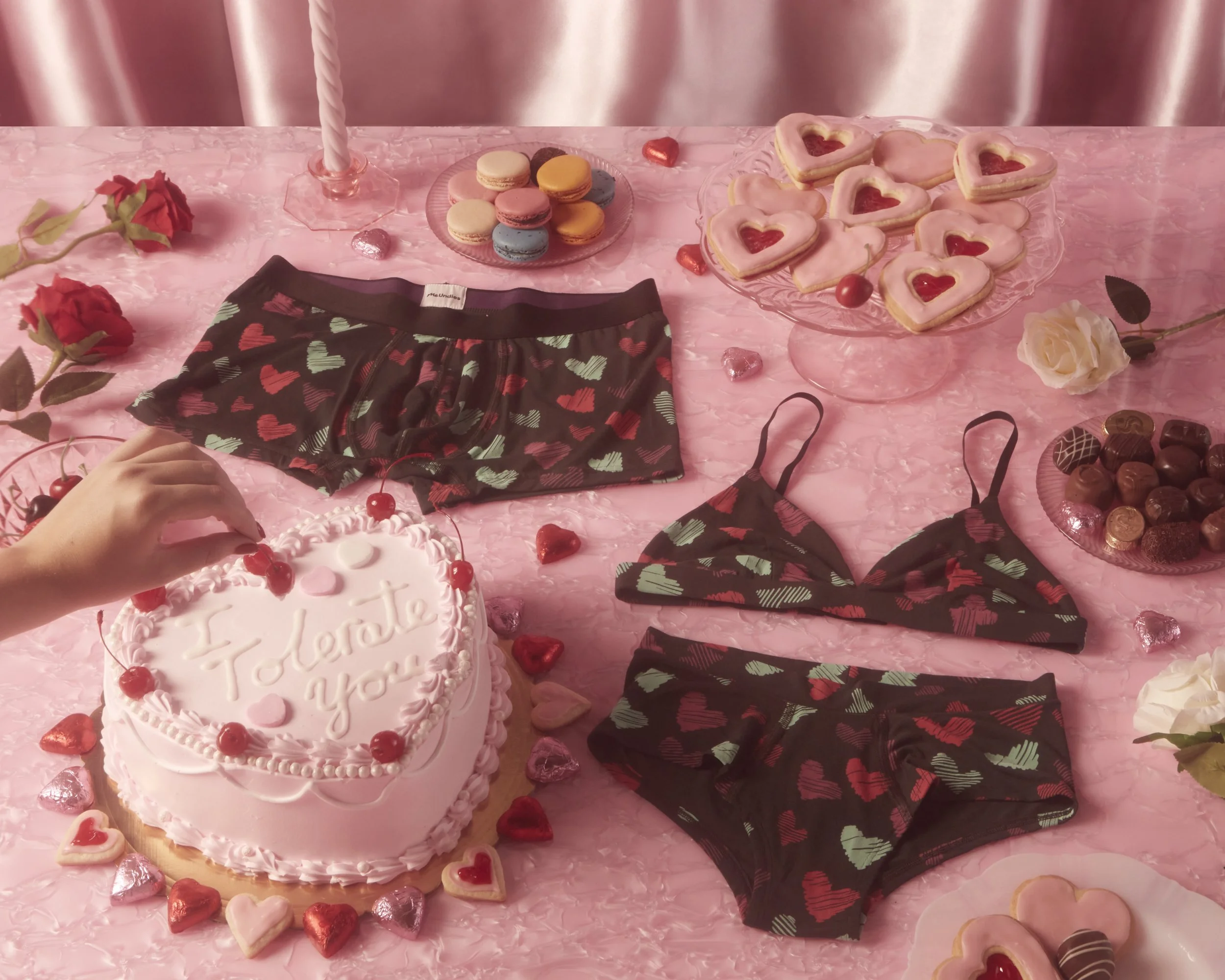 Valentine's Day themed table setup with a heart-shaped cake with a message 'I tolerate you', various heart-shaped cookies, macarons, chocolates, and lingerie, all decorated with roses and pink accents. MeUndies Fashion flatlay photography Los Angeles