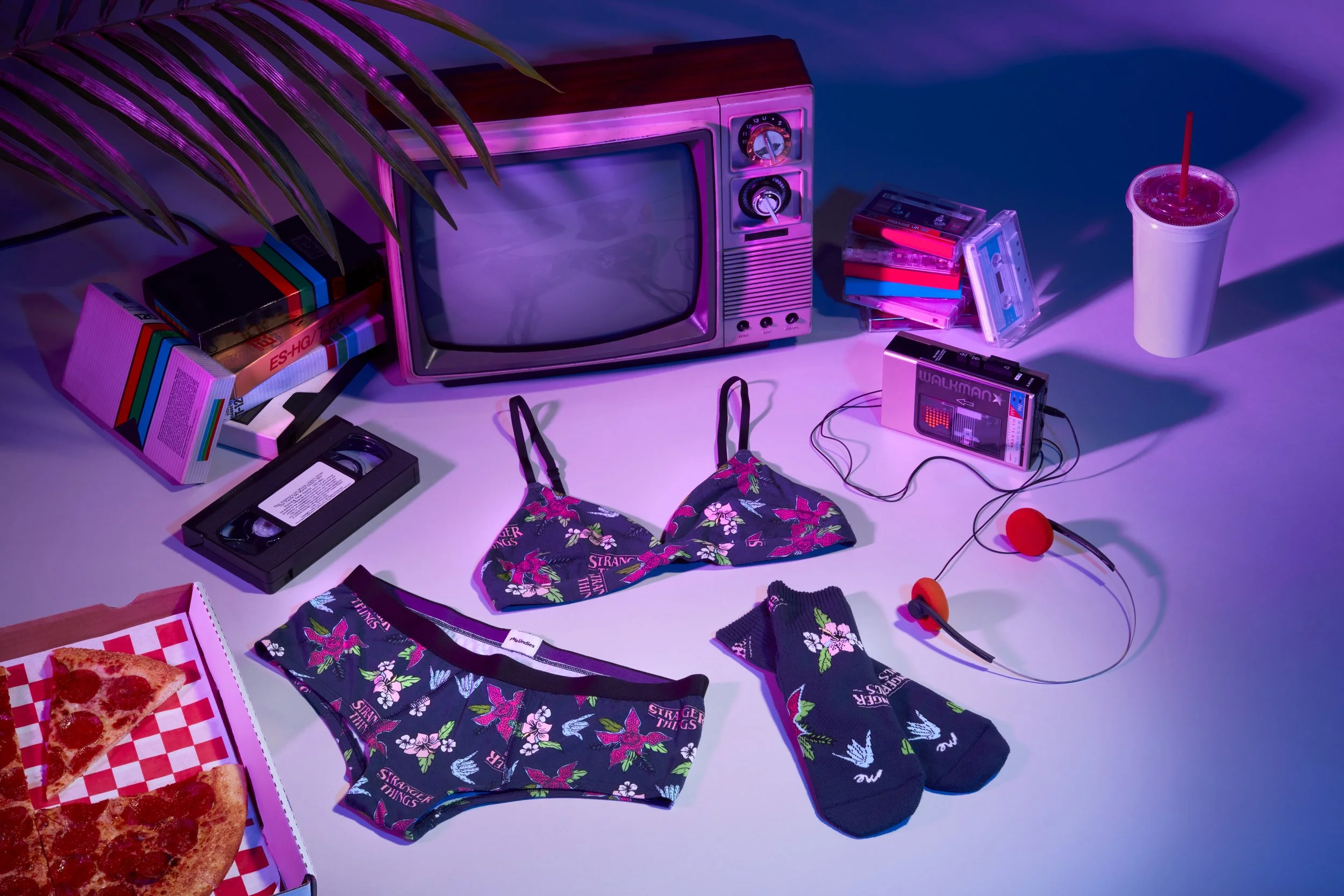 A colorful vintage room scene with a small TV, cassette tapes, a pizza box with slices, floral lingerie and socks, a drink, and a portable gaming device with earbuds, illuminated by purple and pink lighting. MeUndies fashion flatlay photography.