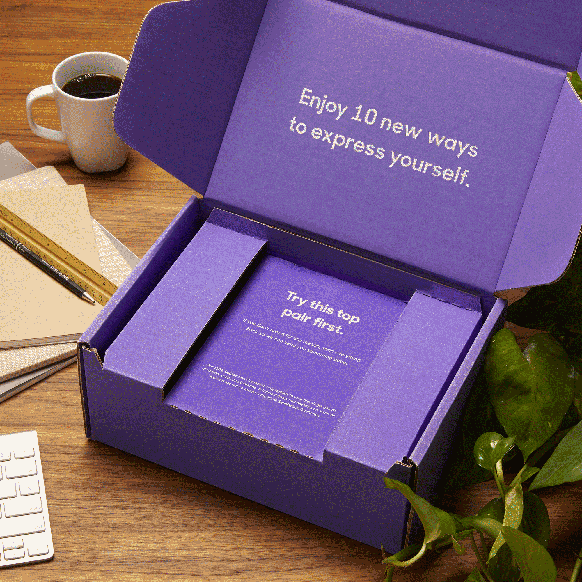 Open a purple box with a GIF video looping, surrounded by a coffee mug, a ruler, notebooks, a pen, part of a keyboard, and green houseplants on a wooden desk. MeUndies fashion flatlay photography Los Angeles.