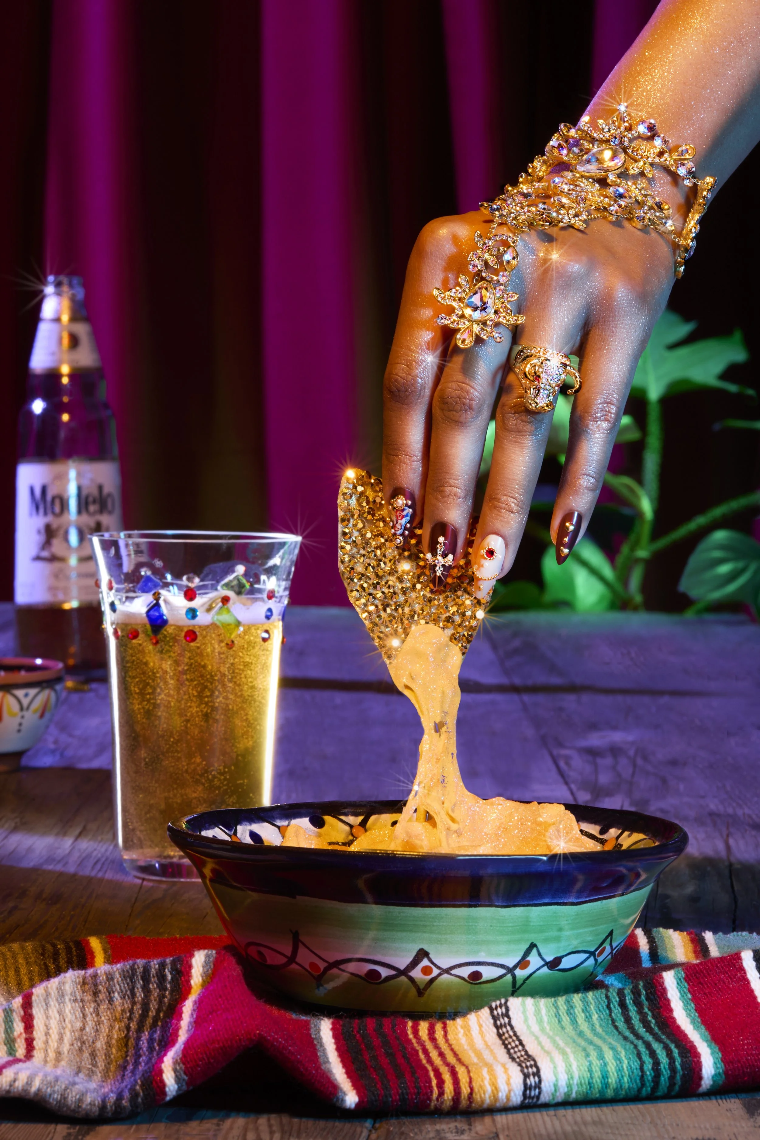 Hand with decorated nails and jewelry reaching into a bowl of cheesy nachos at a festive setting with a Modelo beer bottle, a glass of bubbly, and a colorful tablecloth. Los Angeles-based food photography.